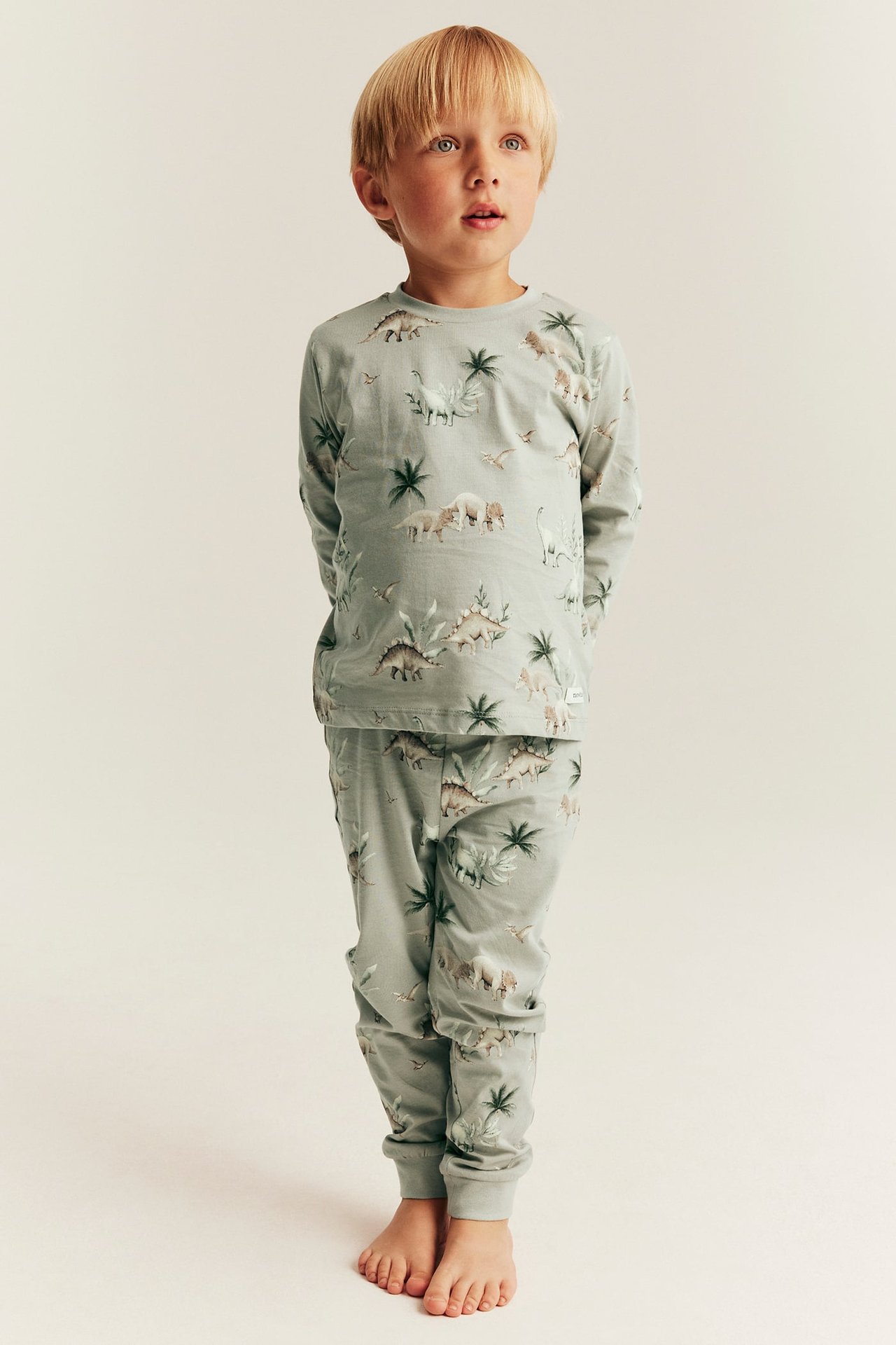 Edestä päin, lapsi yllään kaksiosainen vaaleanvihreä puuvillapyjama dinosauruskuviolla.
