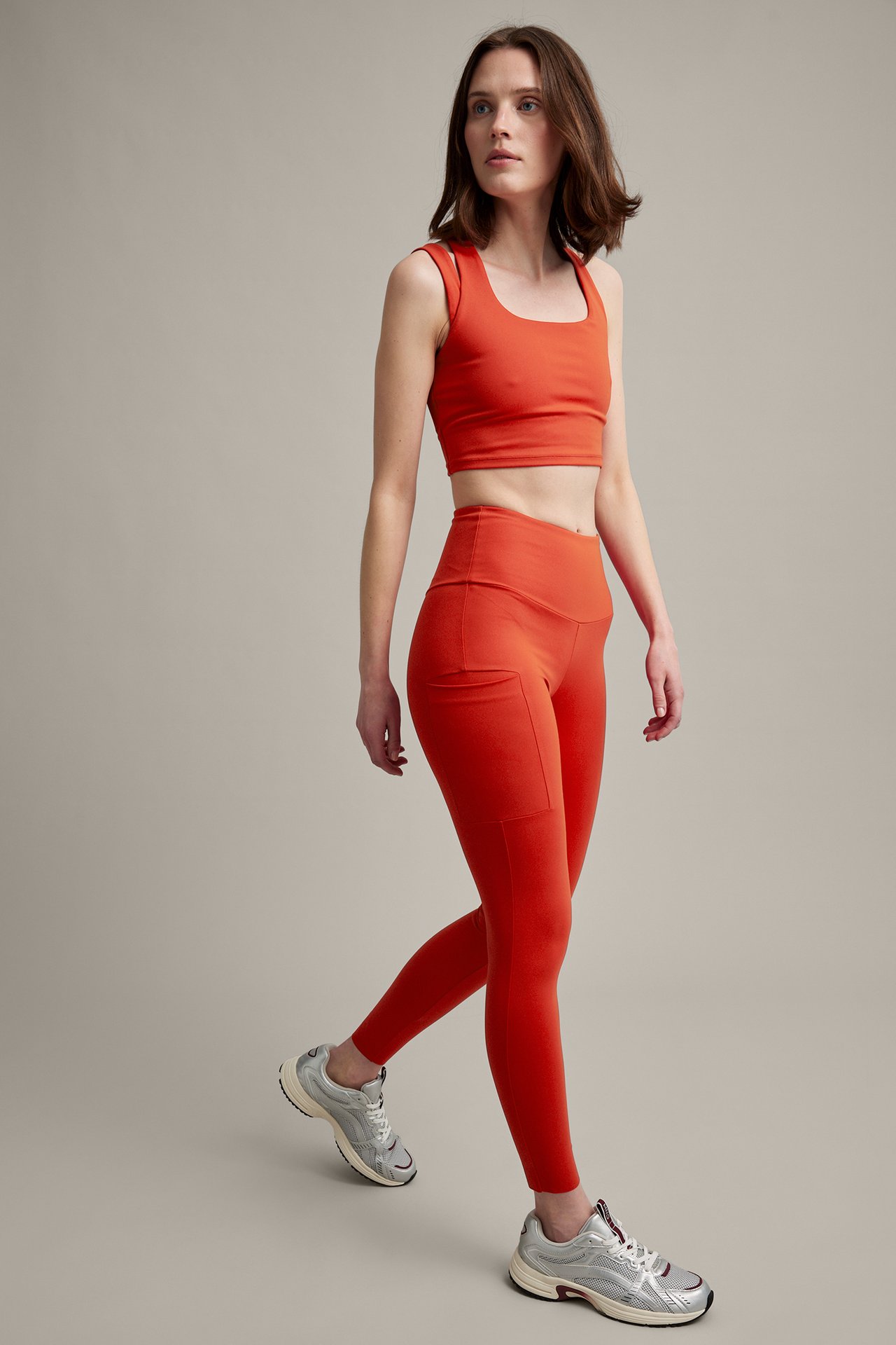 Edestä, naismalli oranssissa crop topissa, taskullisissa leggingsseissä ja hopealenkkareissa.