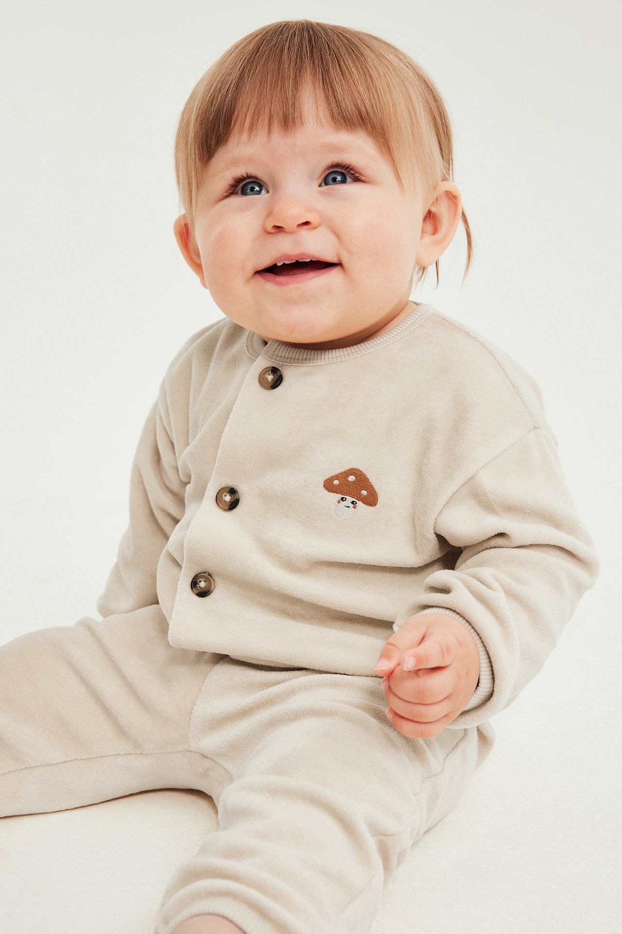 Frontbilde av baby i beige kneppet cardigan med soppbroderi og matchende bukser.
