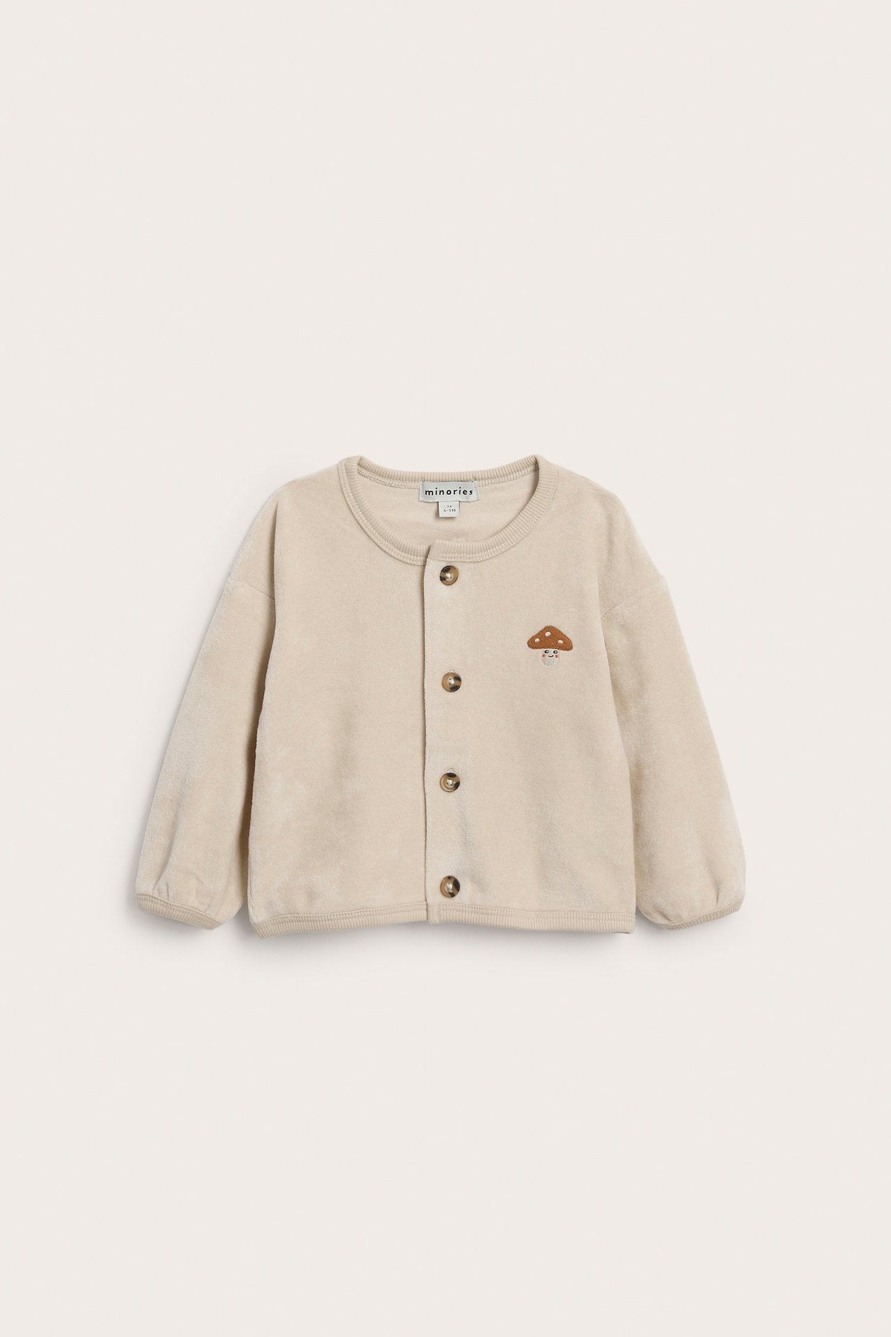 Forfra av lys beige babyjakke med soppbroderi og fem brune knapper.