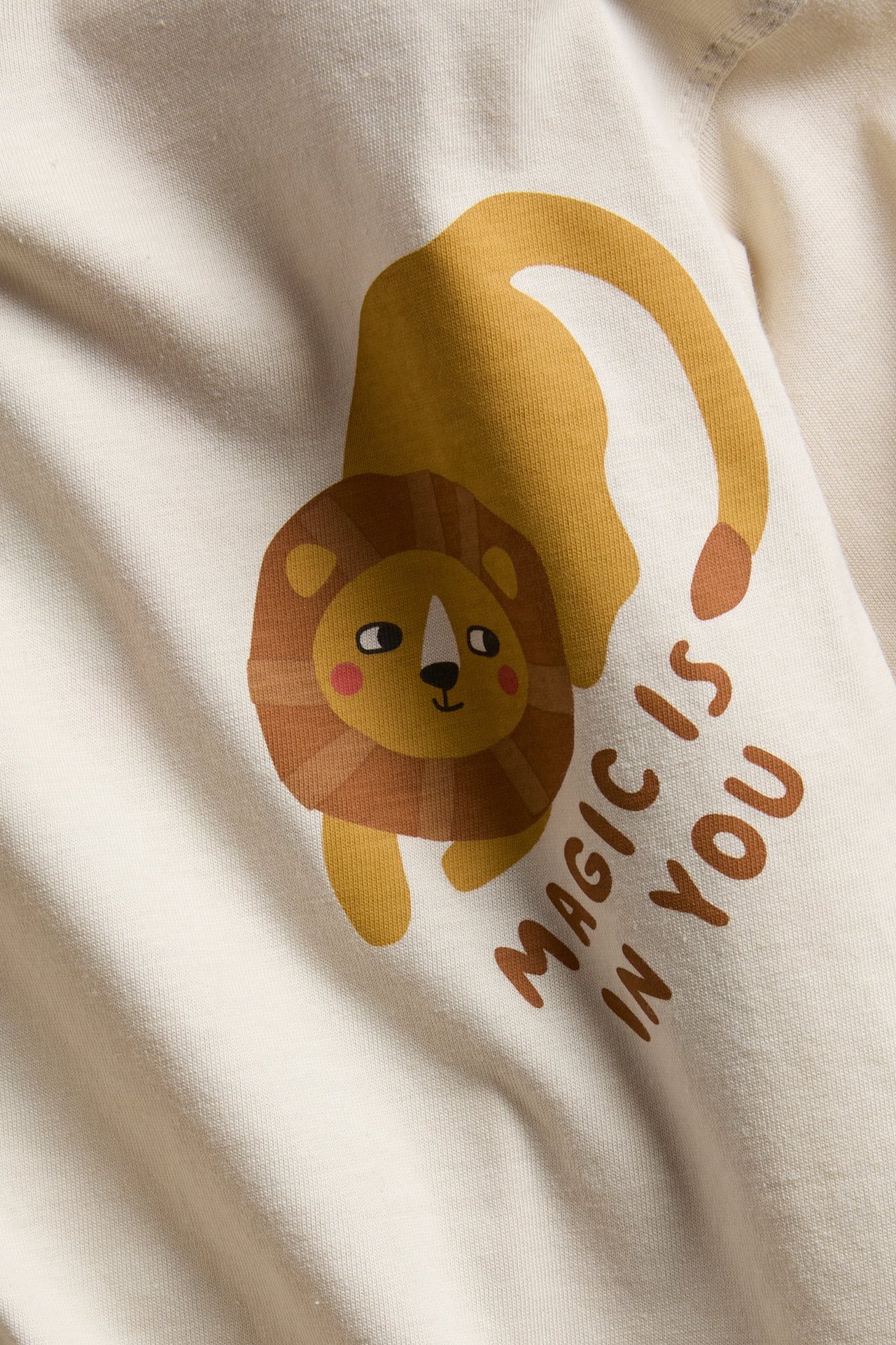 Zbliżenie na kremowe ubranie dziecięce z rysunkowym lwem i napisem „MAGIC IS IN YOU”.