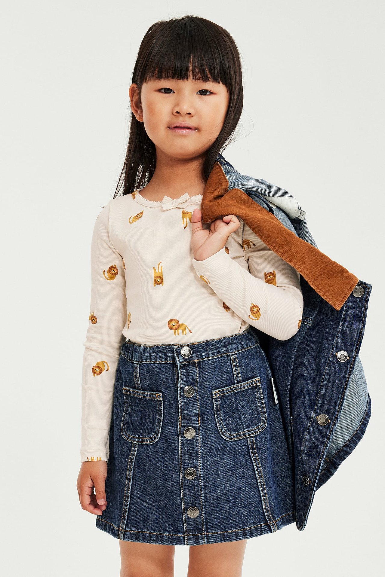 Barn i blått denimknappskjørt, beige langermet topp med løvemønster og denimjakke.