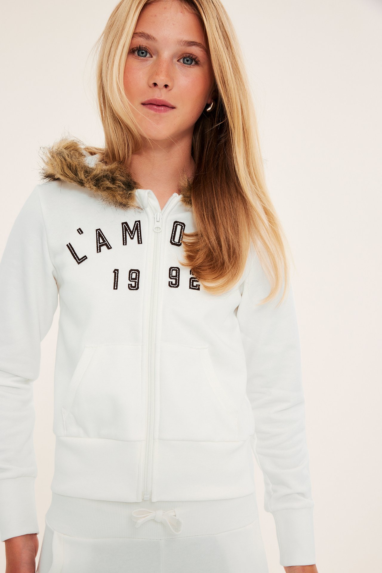 Frontvy av ett barn i vit hoodie med dragkedja, pälsfodrad luva och matchande mjukisbyxor.