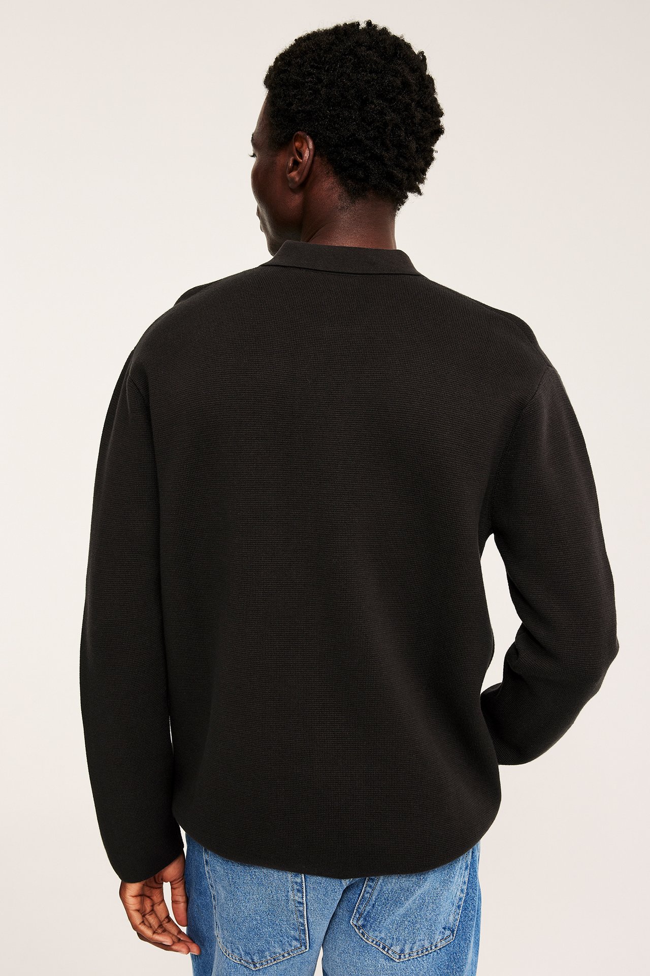 Bakfra av mannlig modell iført en svart teksturert langermet pikétrøye og blå jeans.