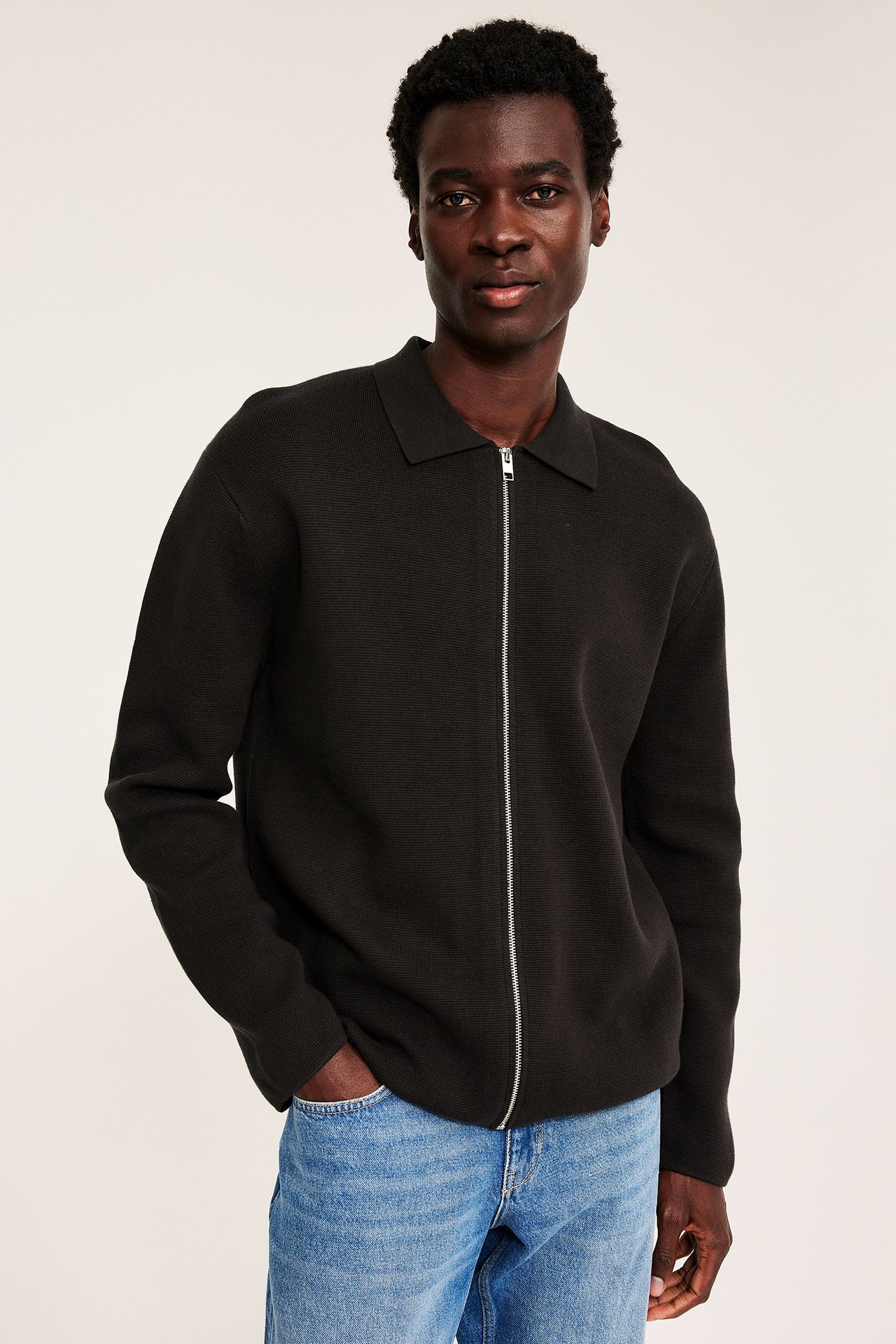 Front, voksen mann i svart hel glidelås ribbestrikket genser og blå jeans.
