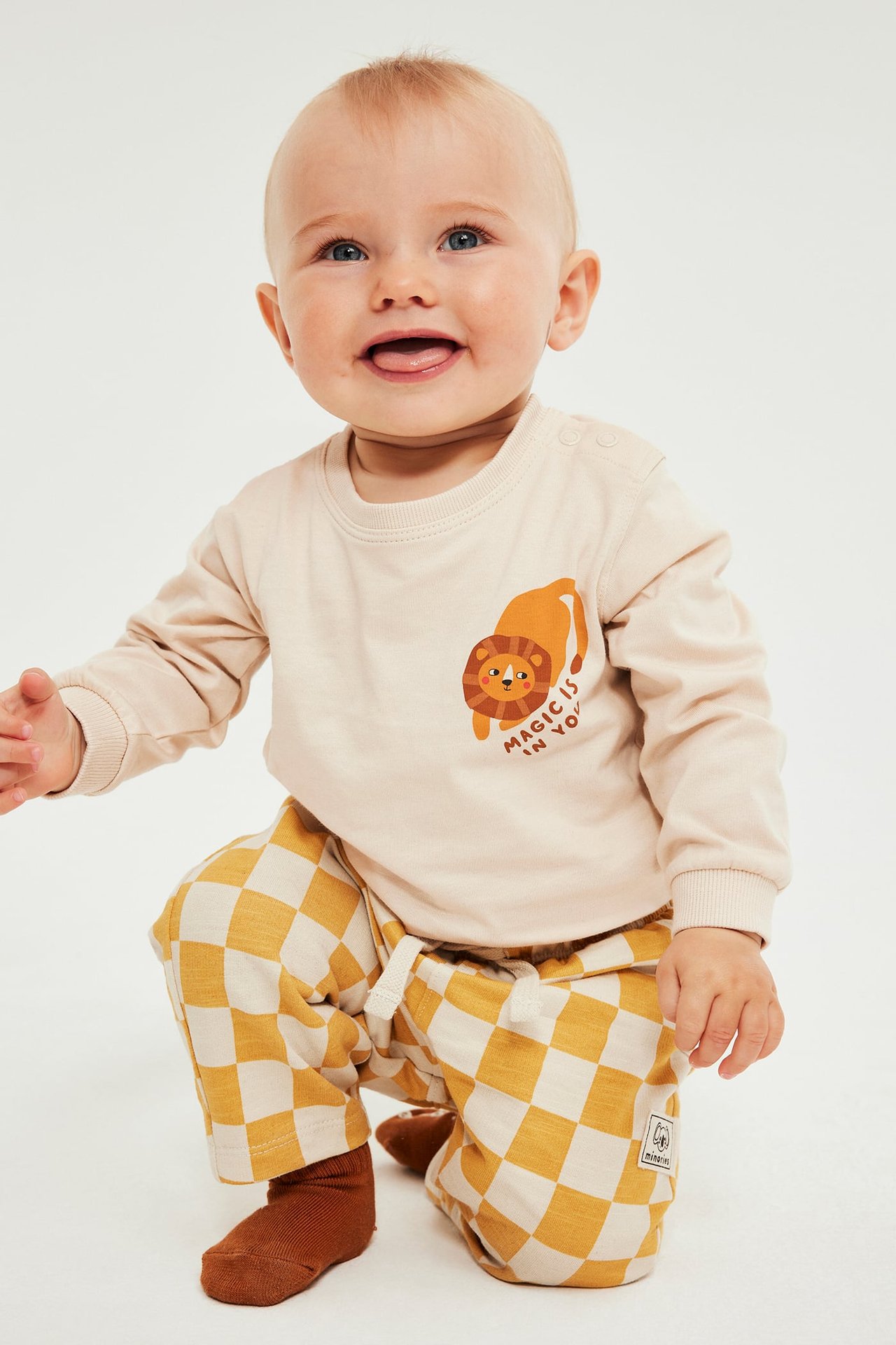 Forfra, baby i beige langermet topp med løvetrykk, gulrutete bukser og brune sokker.