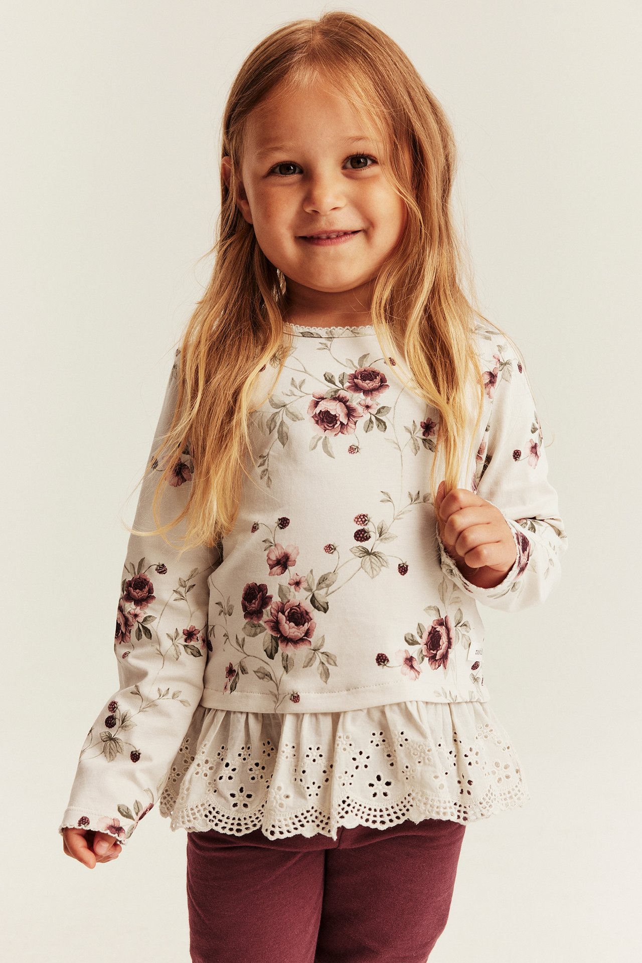 Frontalvisning av et barn iført blomstret peplum-langermet topp med blondekant og mørkerøde leggings.