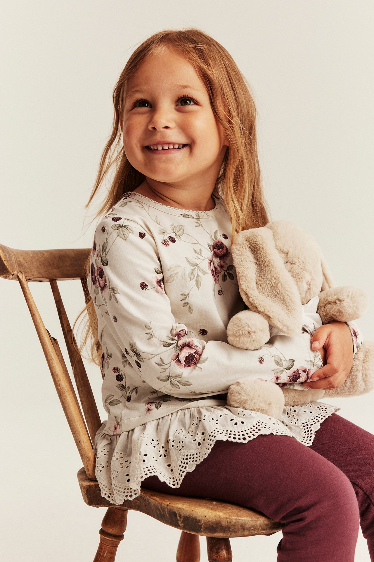 Barns kremhvit blomstret langermet topp med blondekant og burgunder leggings, holder en beige kanin.
