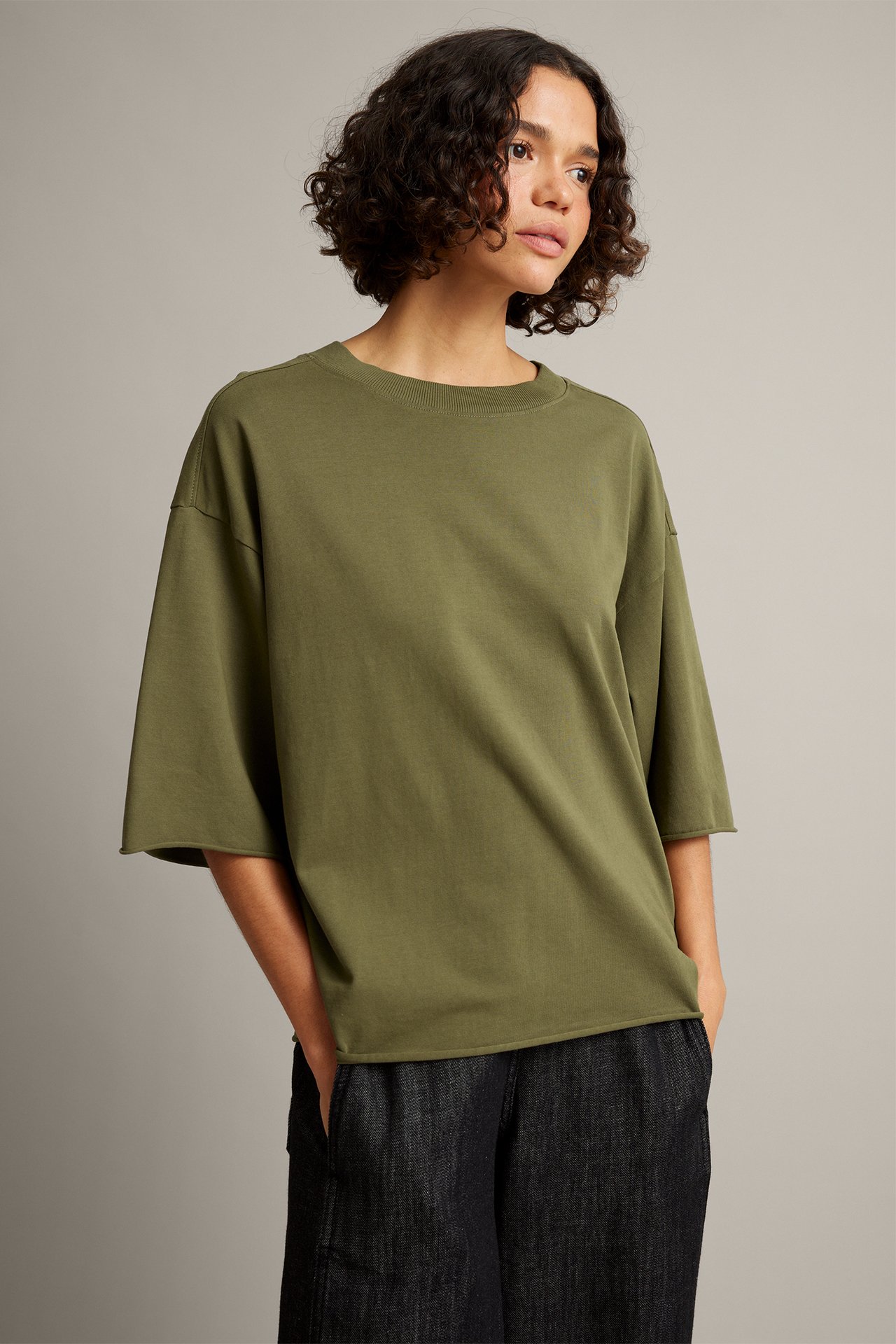 Widok z przodu: kobieta w oversize’owym oliwkowym t-shircie z surowymi brzegami, ciemne dżinsy.