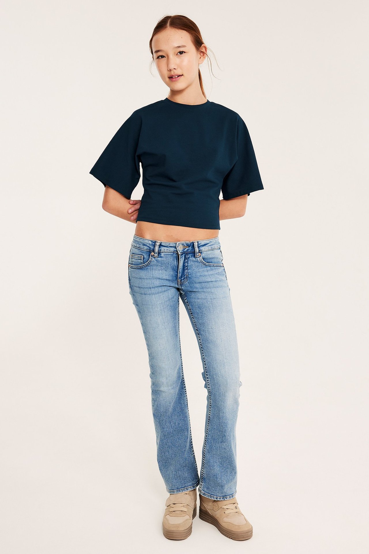 Frontvy av ett barn i en mörkblå kortärmad croppad topp, ljusblå jeans och beiga platåsneakers.