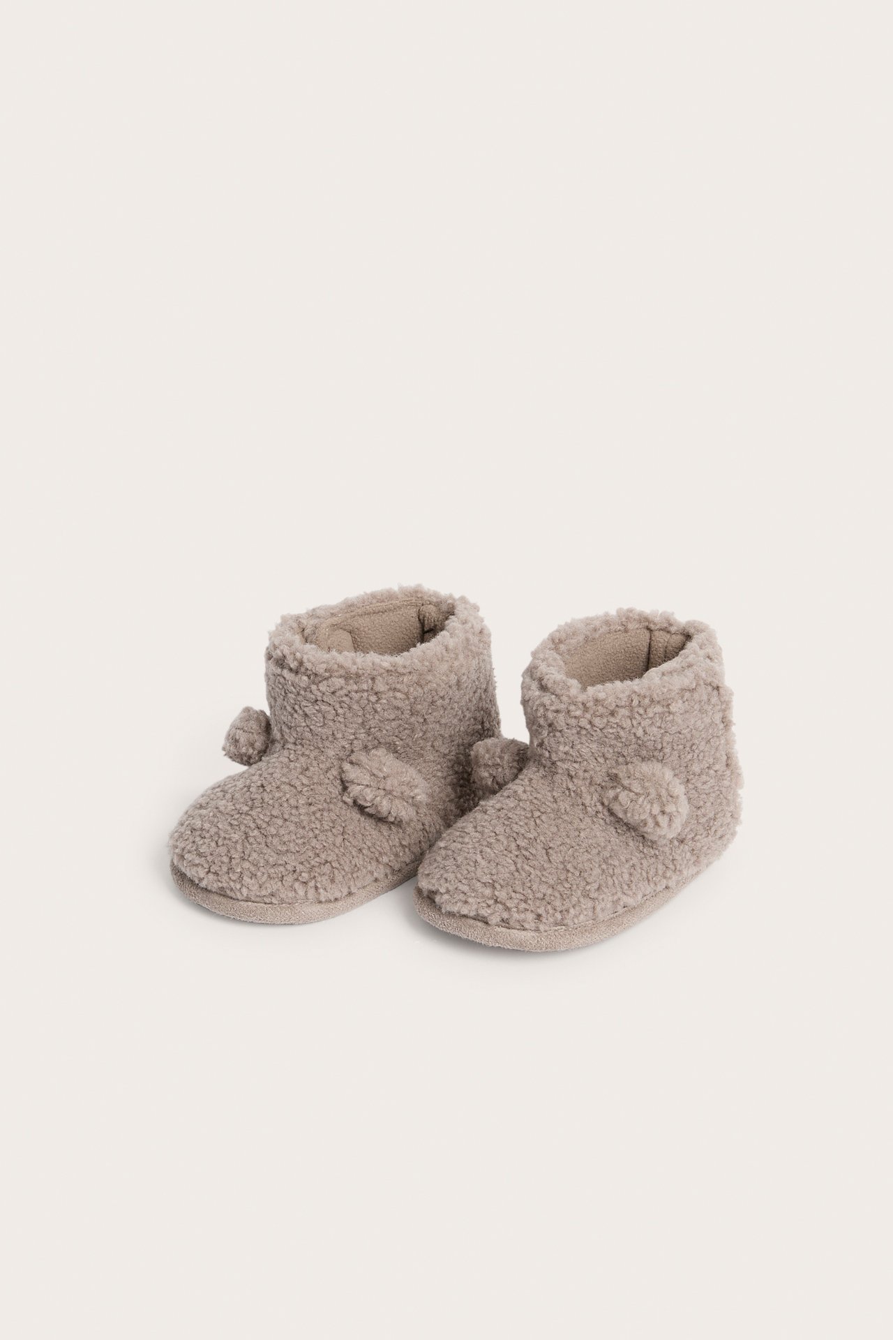 Framifrån, två beiga babytofflor i teddytyg med små dekorativa tofsar.