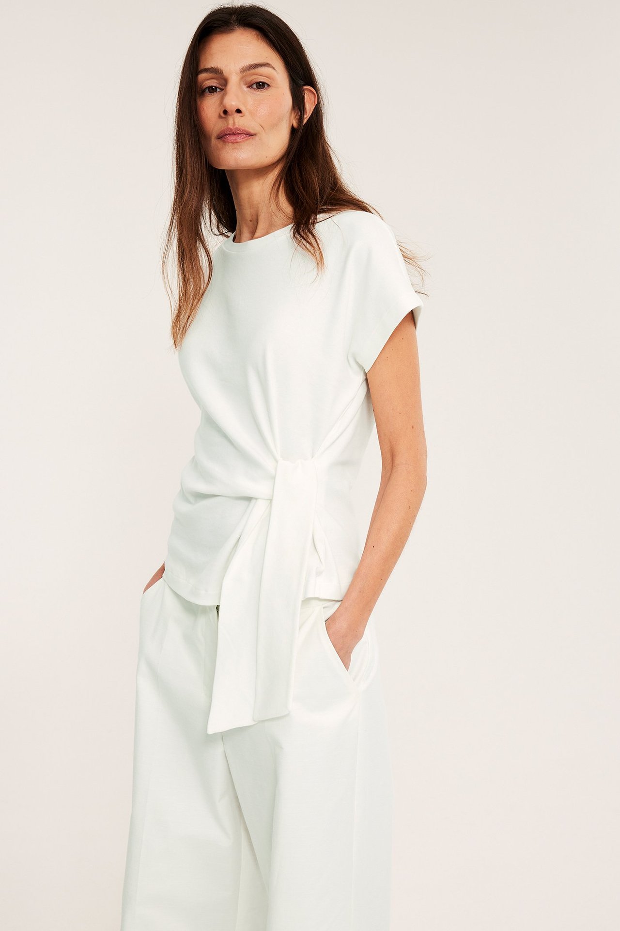 Framifrån, kvinna i en offwhite kortärmad bomullstopp med knytdetalj, samt matchande offwhite byxor.