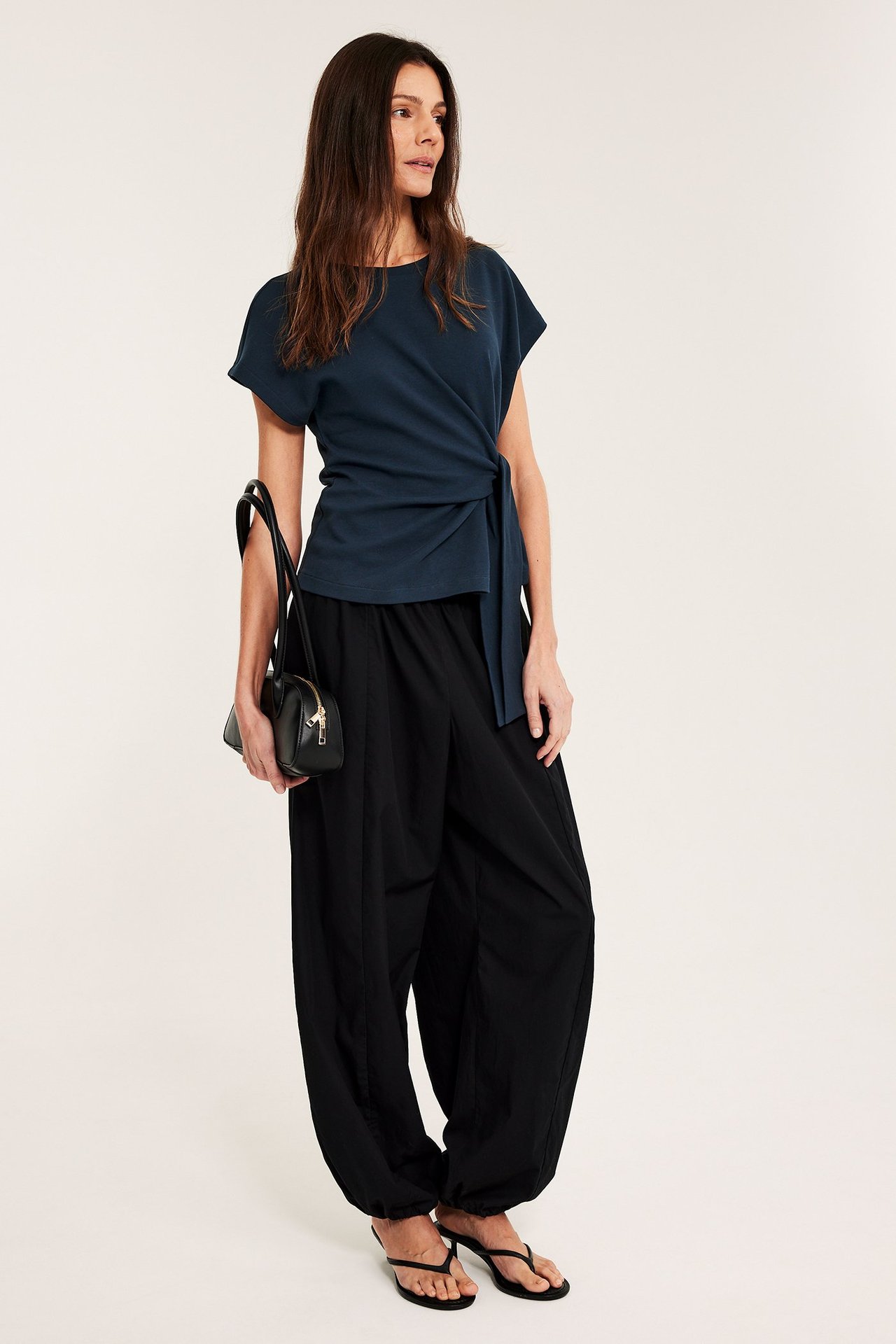 Kobieta w granatowym bawełnianym topie z krótkim rękawem i wiązaniem, czarnych szerokich spodniach, klapkach, trzyma czarną torebkę.