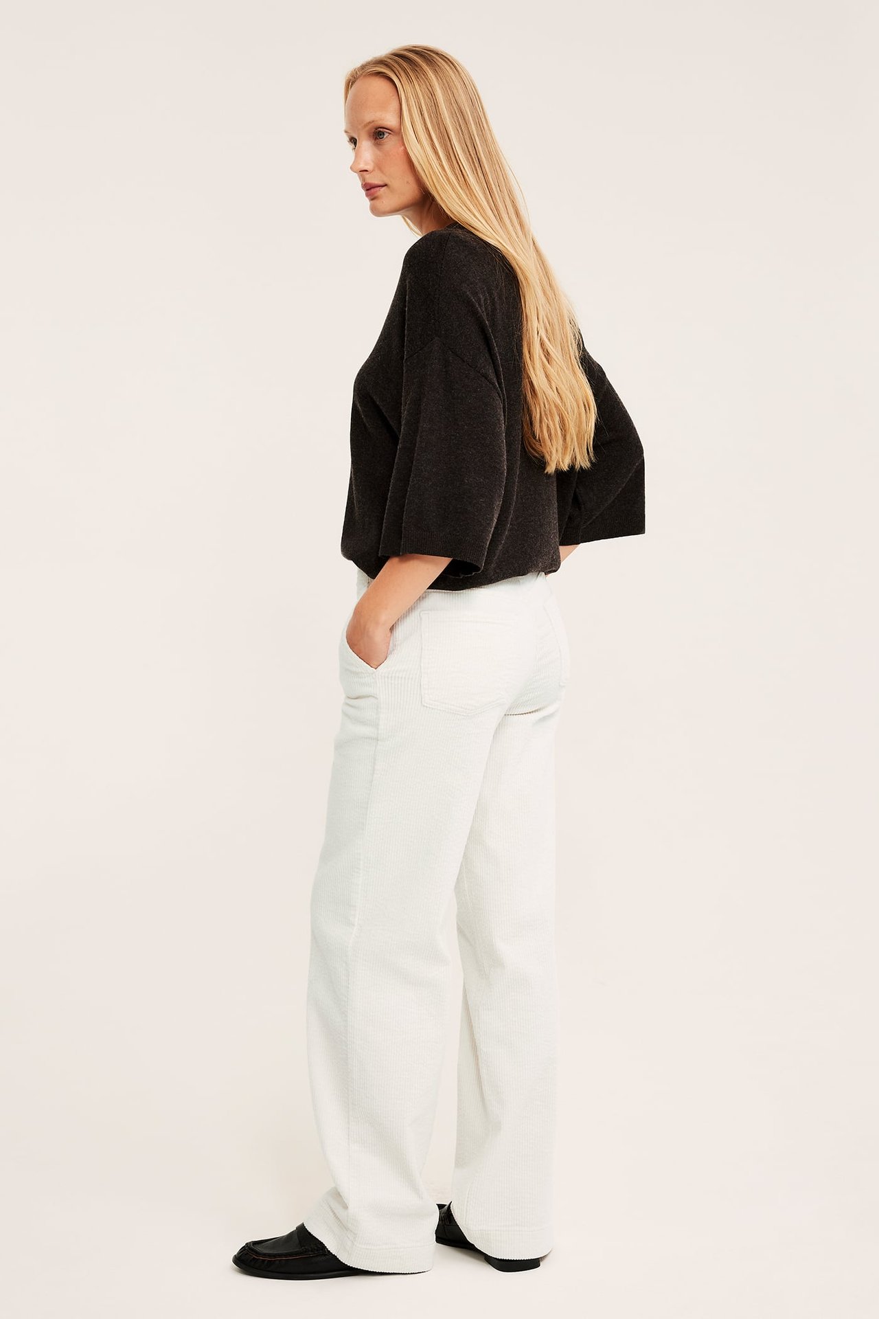 Sidovy av kvinnlig modell i vita vida manchesterbyxor, en mörkgrå oversize topp och svarta loafers.