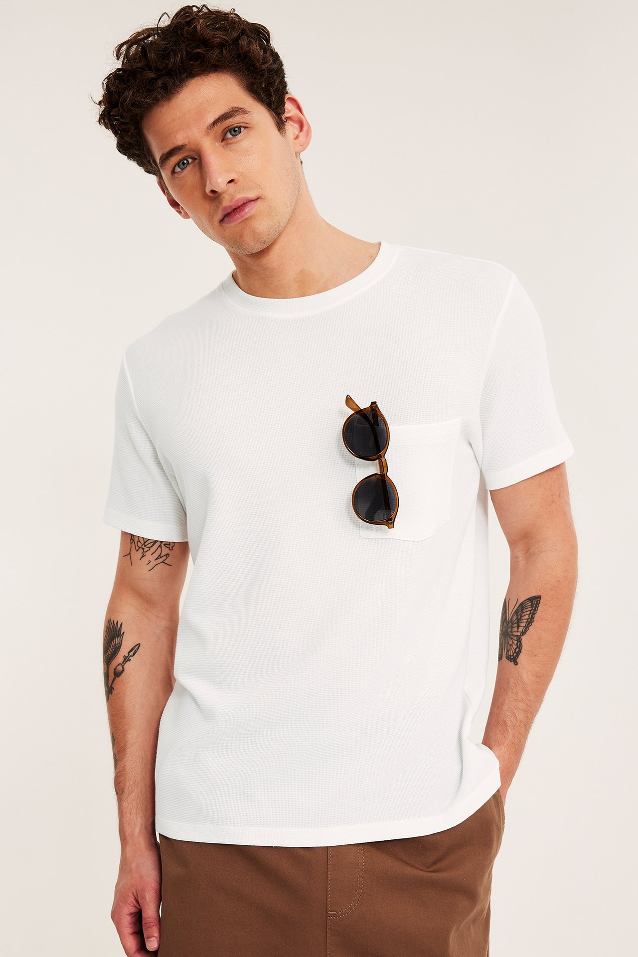 Edestäpäin: mies yllään valkoinen joustinneule-T-paita pyöreällä kaula-aukolla, rintataskussa ruskeat aurinkolasit, ja ruskeat housut.