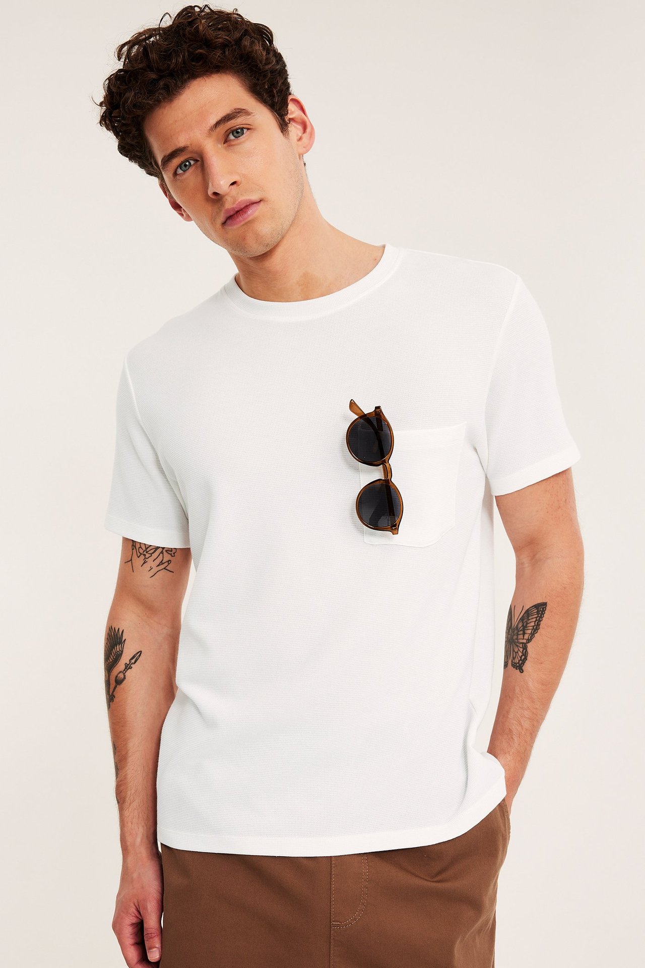Frontbilde: mann i hvit, ribbestrikket T-skjorte med rund hals, brystlomme, brune solbriller og brune bukser.