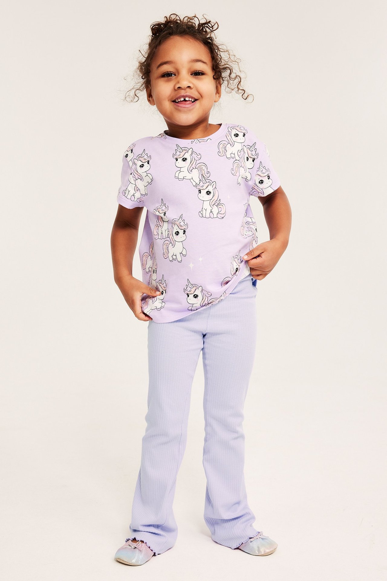 Leende barn, framifrån, bär ljuslila ribbade utsvängda leggings och en lila t-shirt med enhörningsmönster.