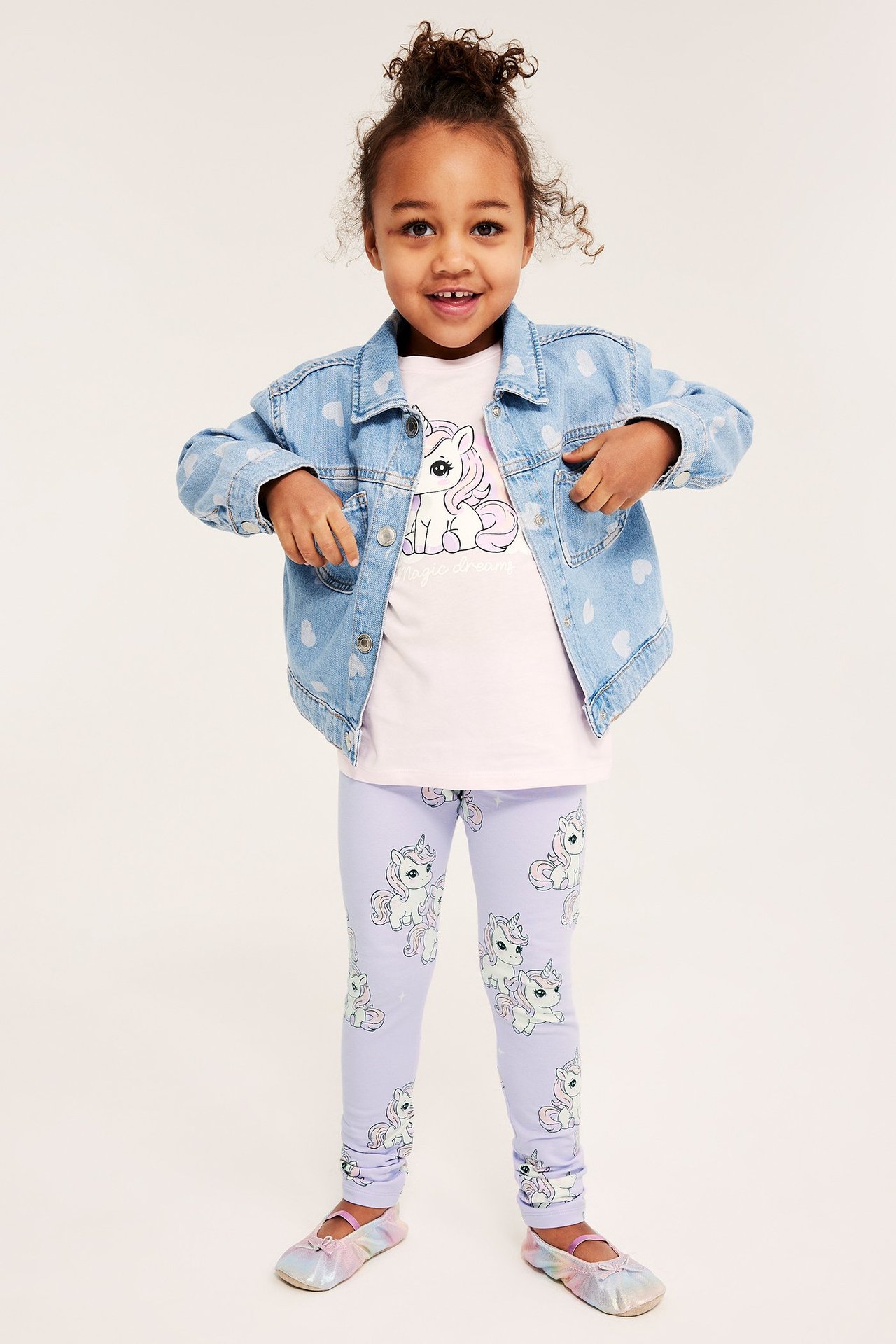 Dziecko ma na sobie liliowe legginsy w jednorożce, różowy t-shirt z jednorożcem, niebieską kurtkę jeansową w serca i kolorowe balerinki.