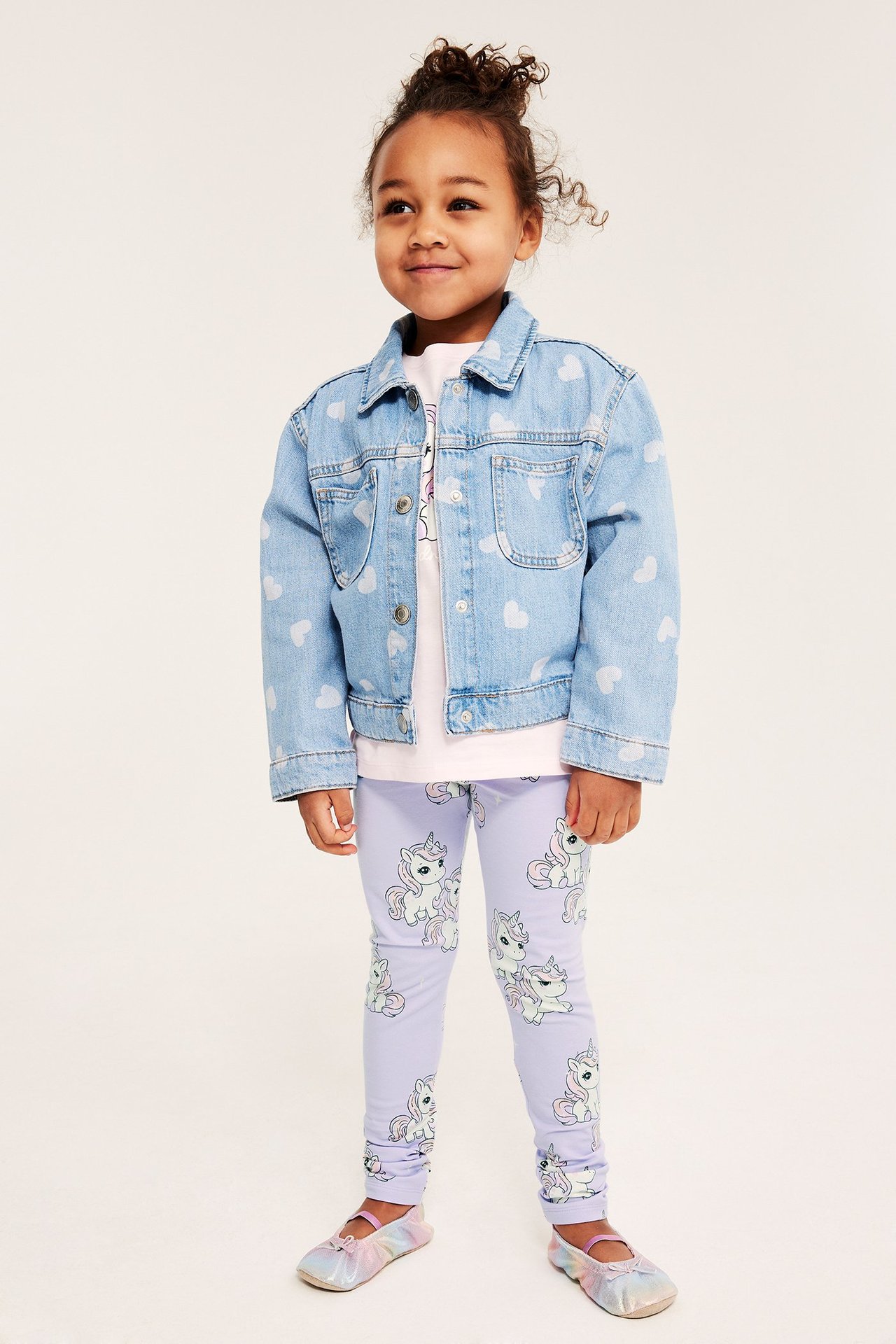 Widok z przodu: Dziecko w jasnofioletowych legginsach w jednorożce, niebieskiej kurtce w serca, różowym T-shircie, opalizujących butach.