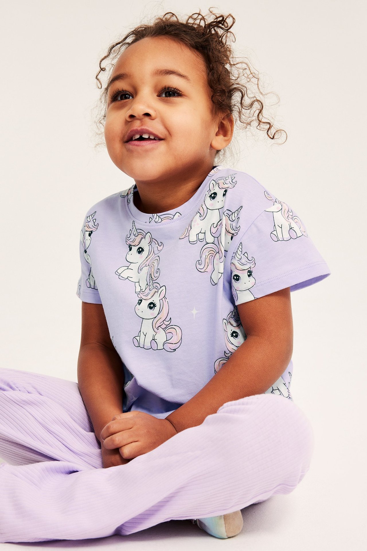 Frontvy av ett barn i ljuslila bomullstopp med enhörningsprint och ljuslila ribbade byxor.