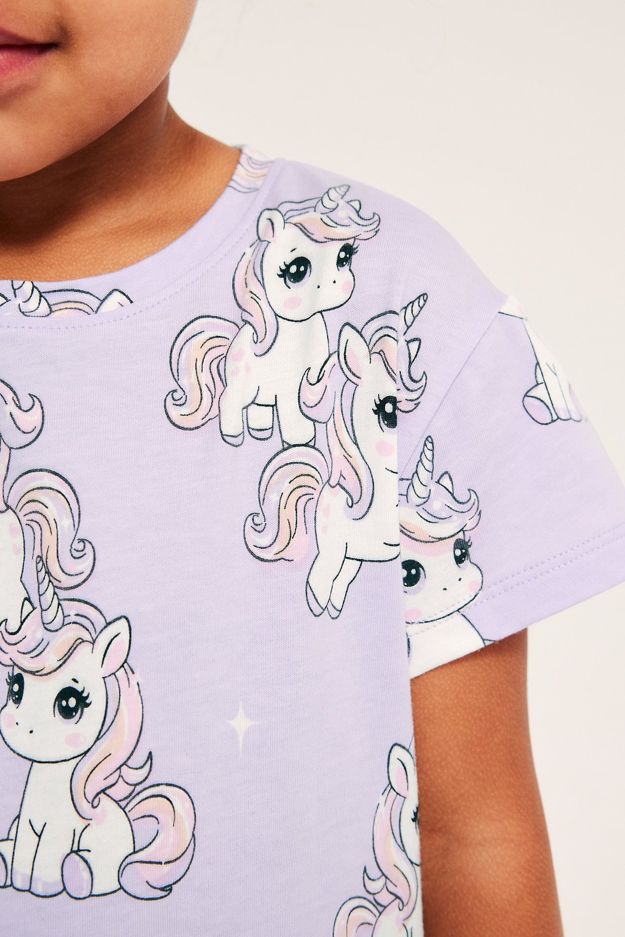 Närbild, framifrån, på en barnets ljuslila bomullst-shirt med tryck av söta enhörningar.