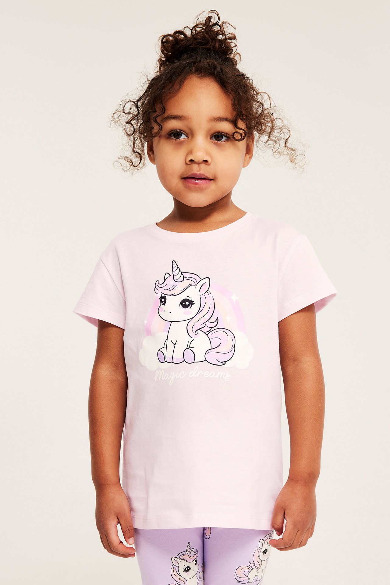 Framifrån, ett barn bär en ljusrosa bomulls-T-shirt med enhörningstryck och lila leggings.