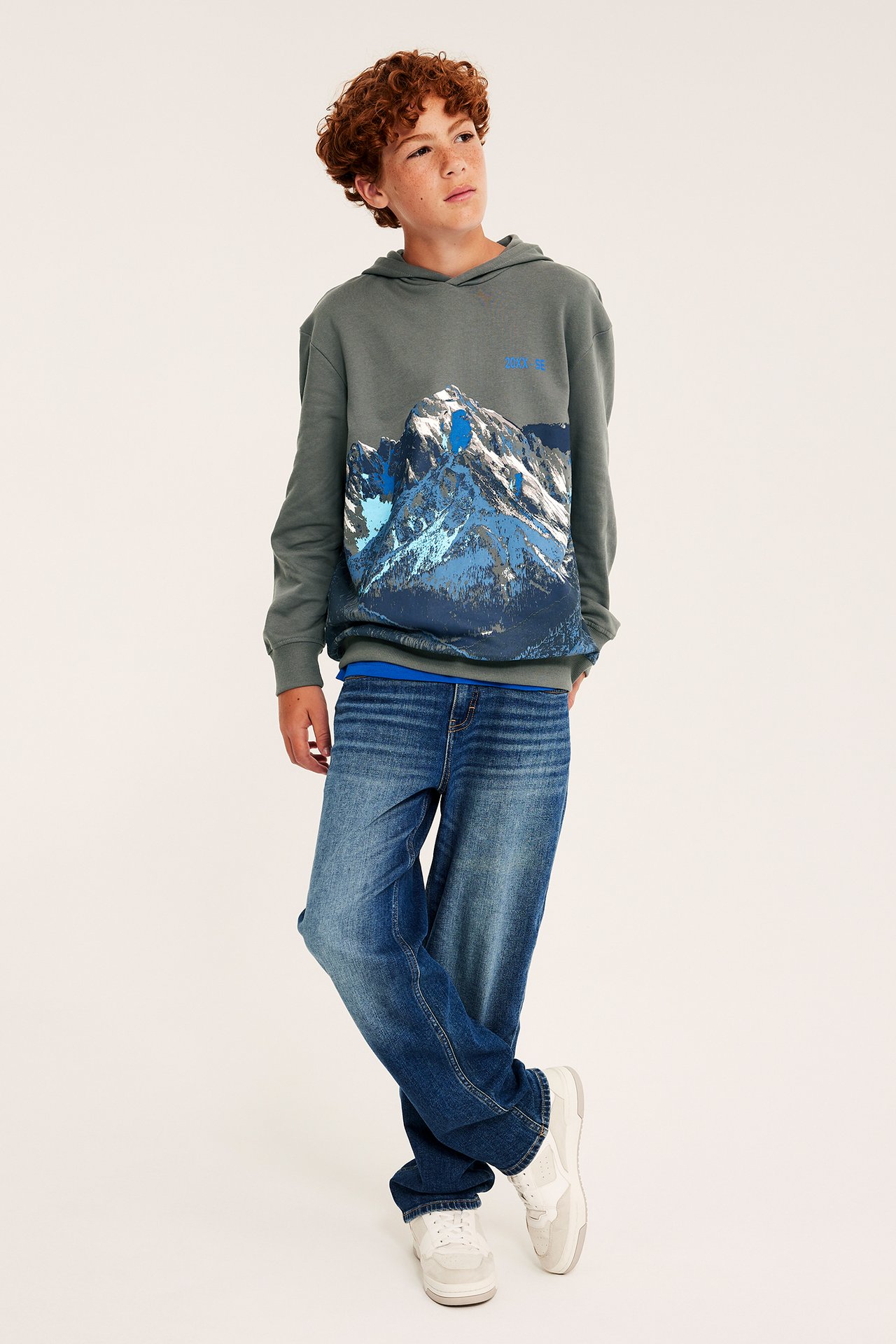 Framifrån, ett barn i en grå hoodie med bergstryck, blå jeans och vita sneakers.