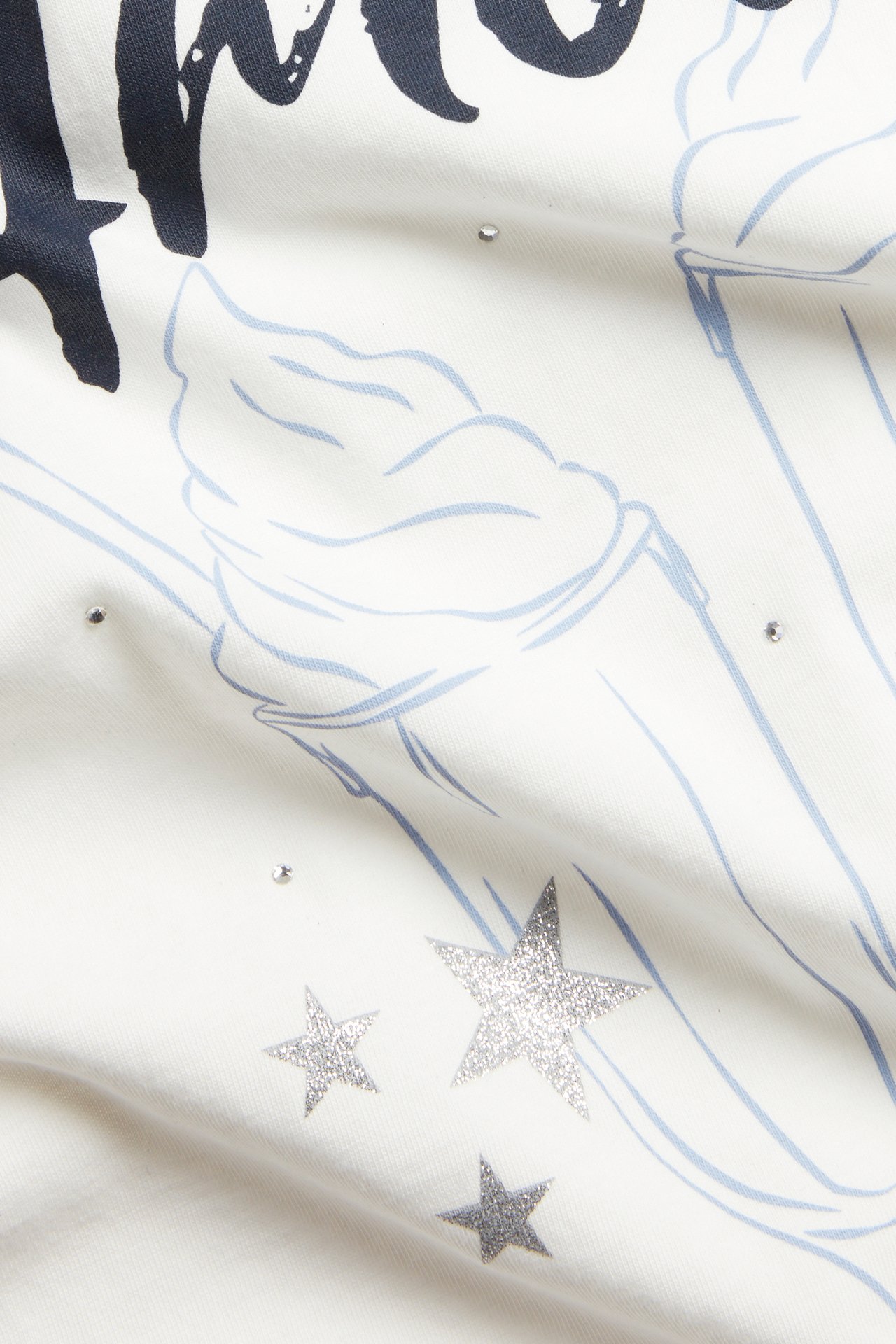 Zbliżenie na bawełniany T-shirt w kolorze ecru z granatowym napisem, jasnoniebieskim rysunkiem i srebrnymi brokatowymi gwiazdkami.