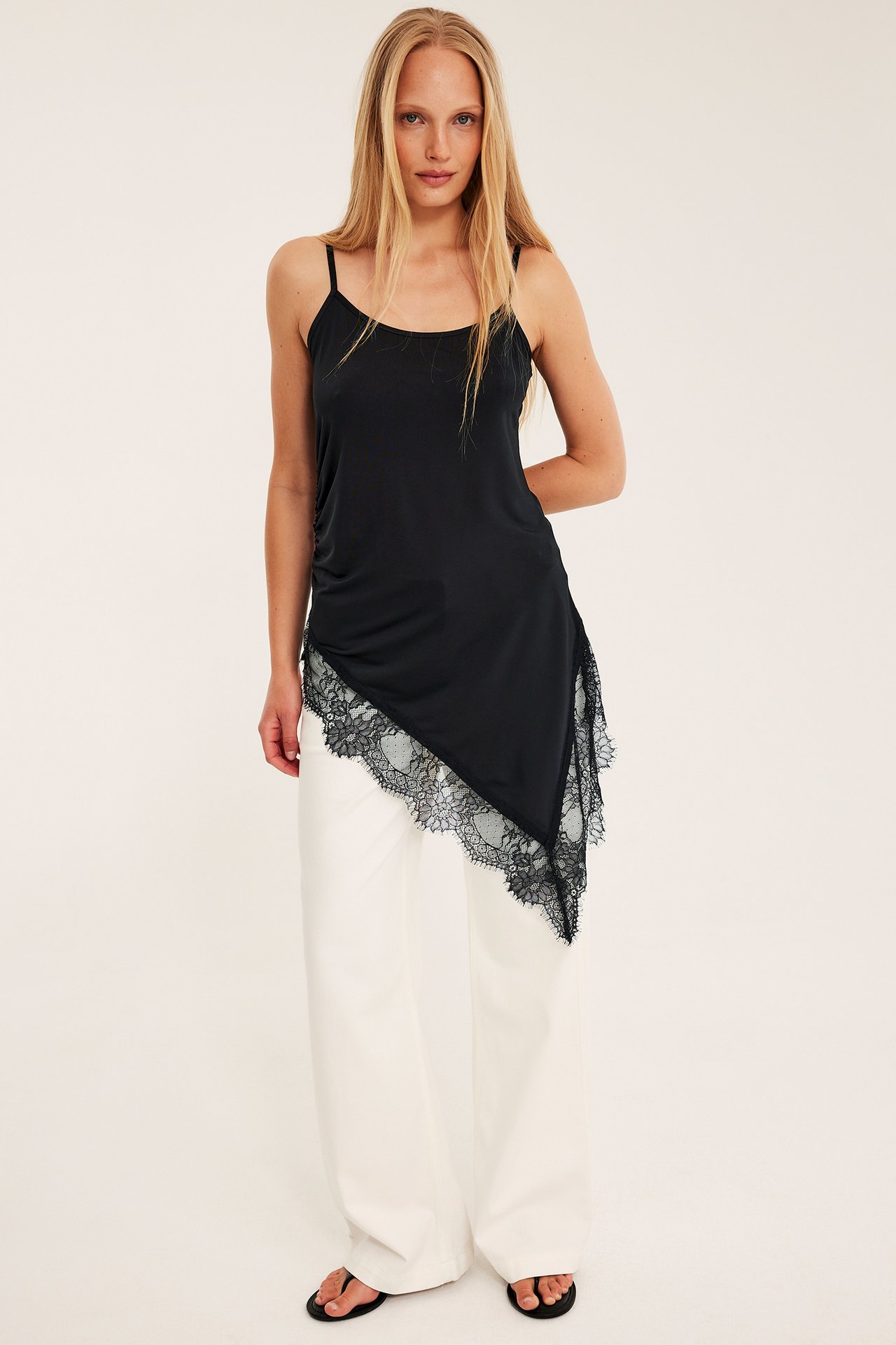 Edestä: Nainen mustassa epäsymmetrisessä pitsitoppauksessa, valkoisissa leveälahkeisissa housuissa ja mustissa sandaaleissa.