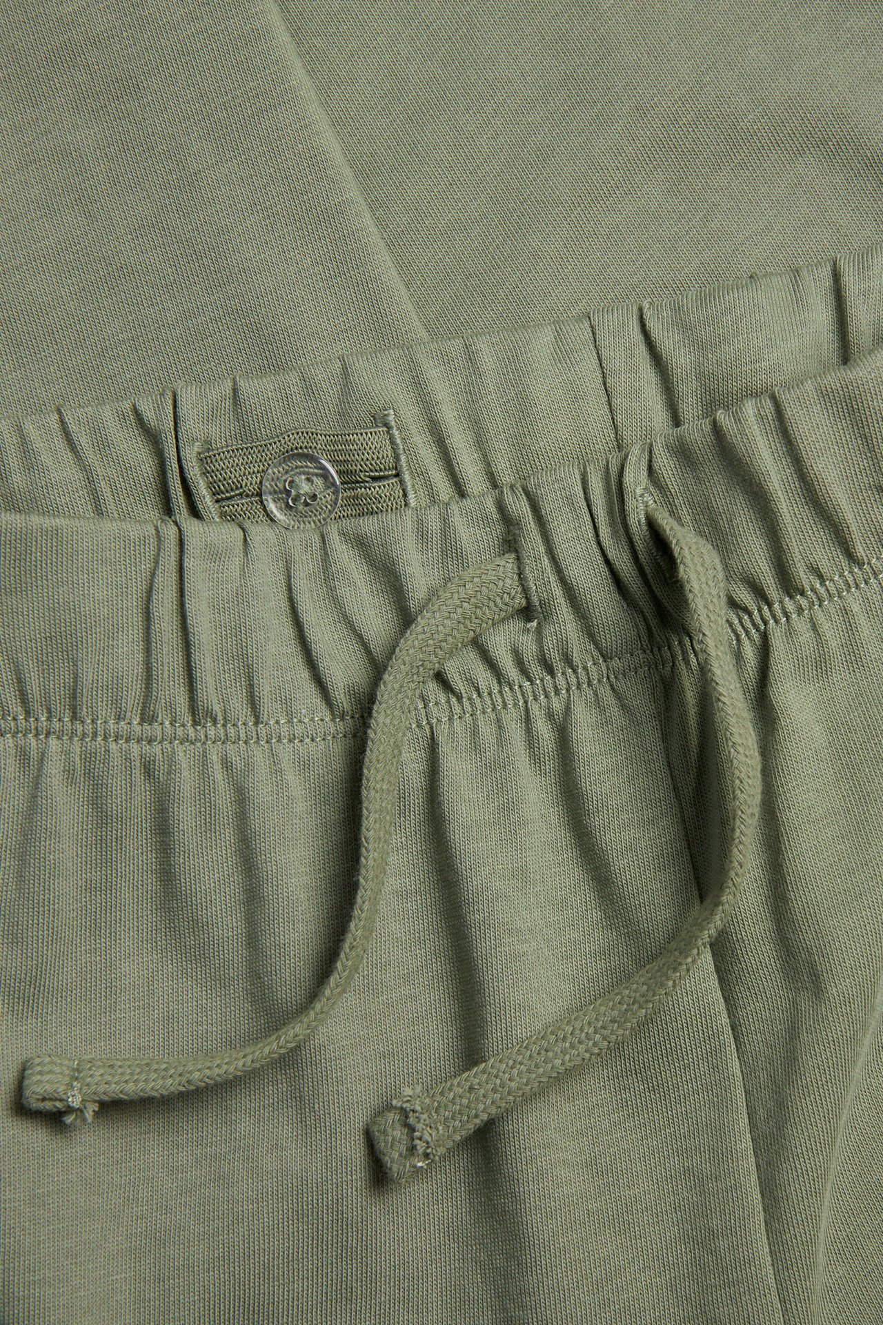 Lähikuva lasten tummanvihreistä puuvilla collegehousuista, joissa joustovyötärö, kiristysnyöri ja säädettävä nappi.