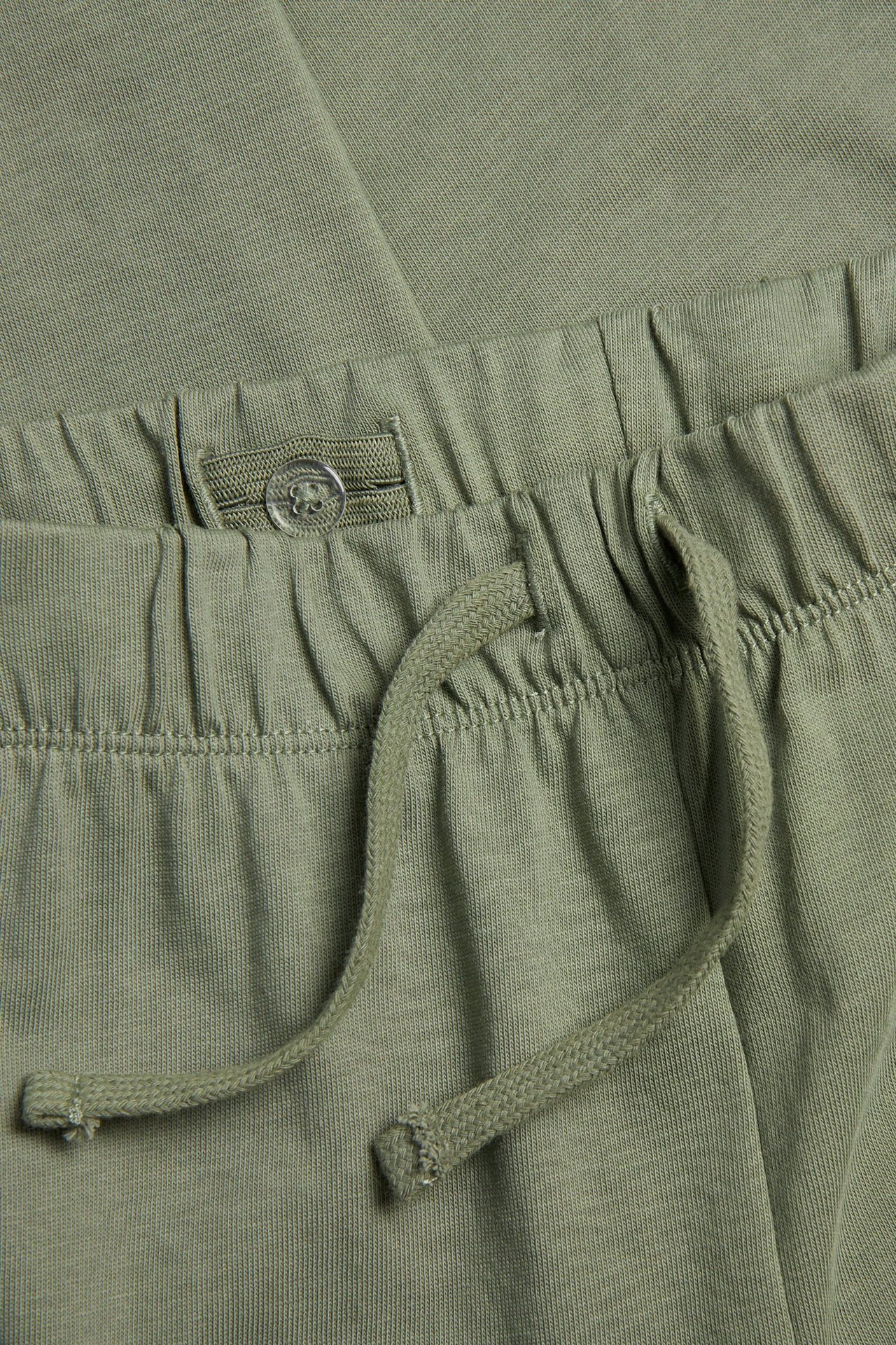 Lähikuva lasten tummanvihreistä puuvilla collegehousuista, joissa joustovyötärö, kiristysnyöri ja säädettävä nappi.