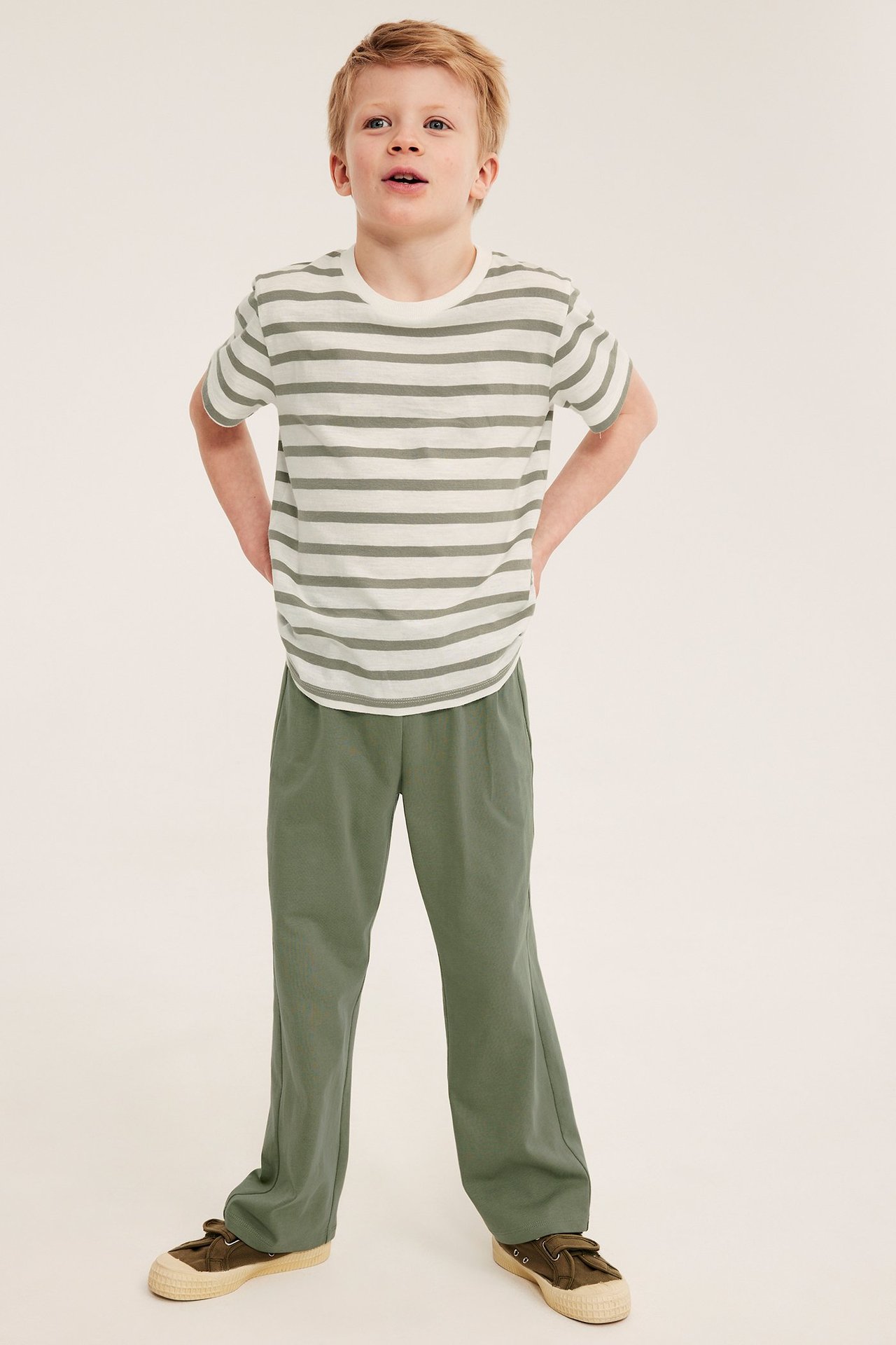 Edestä kuvattu lapsi tummanvihreissä suoralahkeisissa collegehousuissa ja raidallisessa t-paidassa.