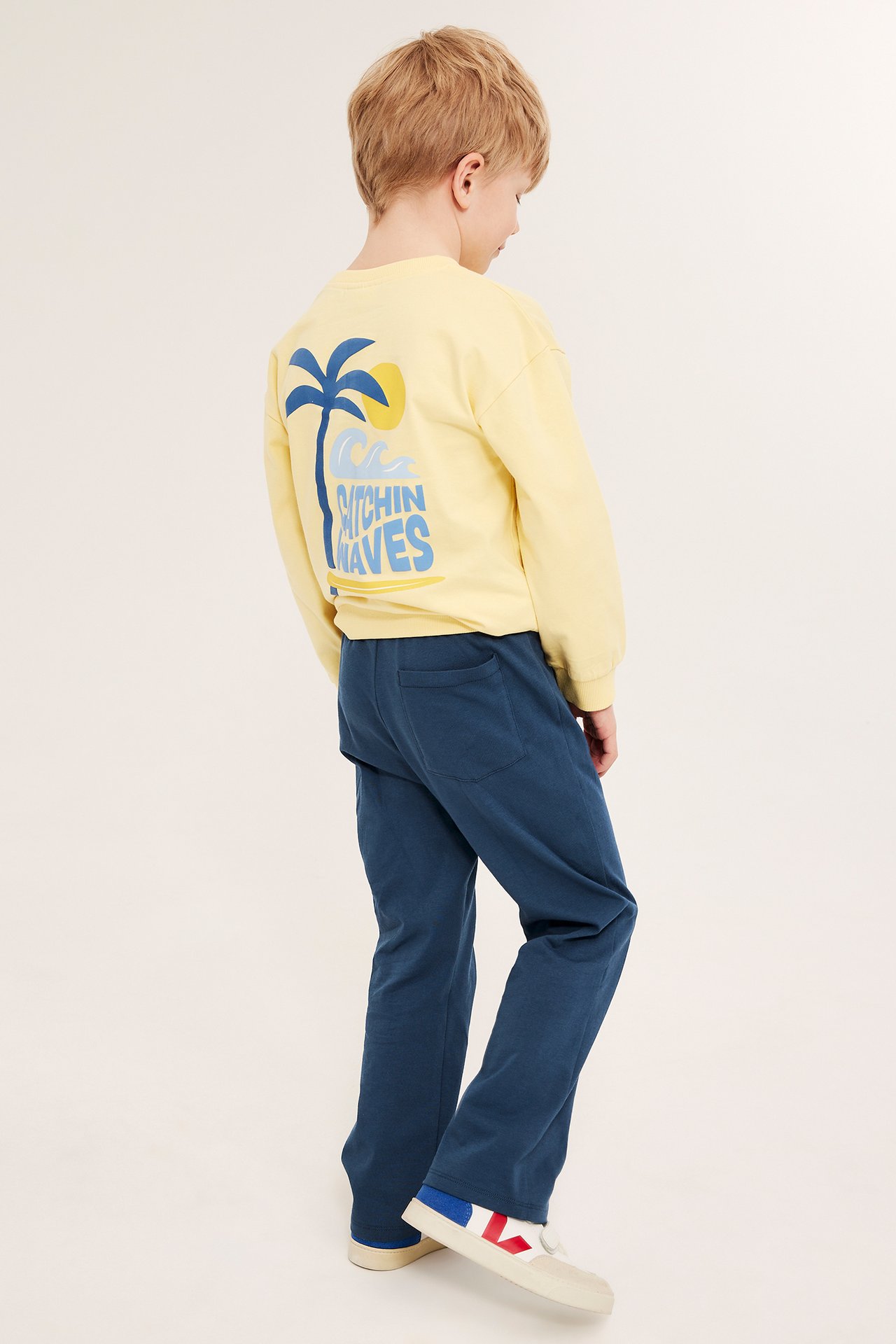 Lapsi takaa päin sinisissä suoralahkeisissa puuvillacollegehousuissa, keltaisessa graafisessa paidassa ja beigenvärisissä tennareissa.