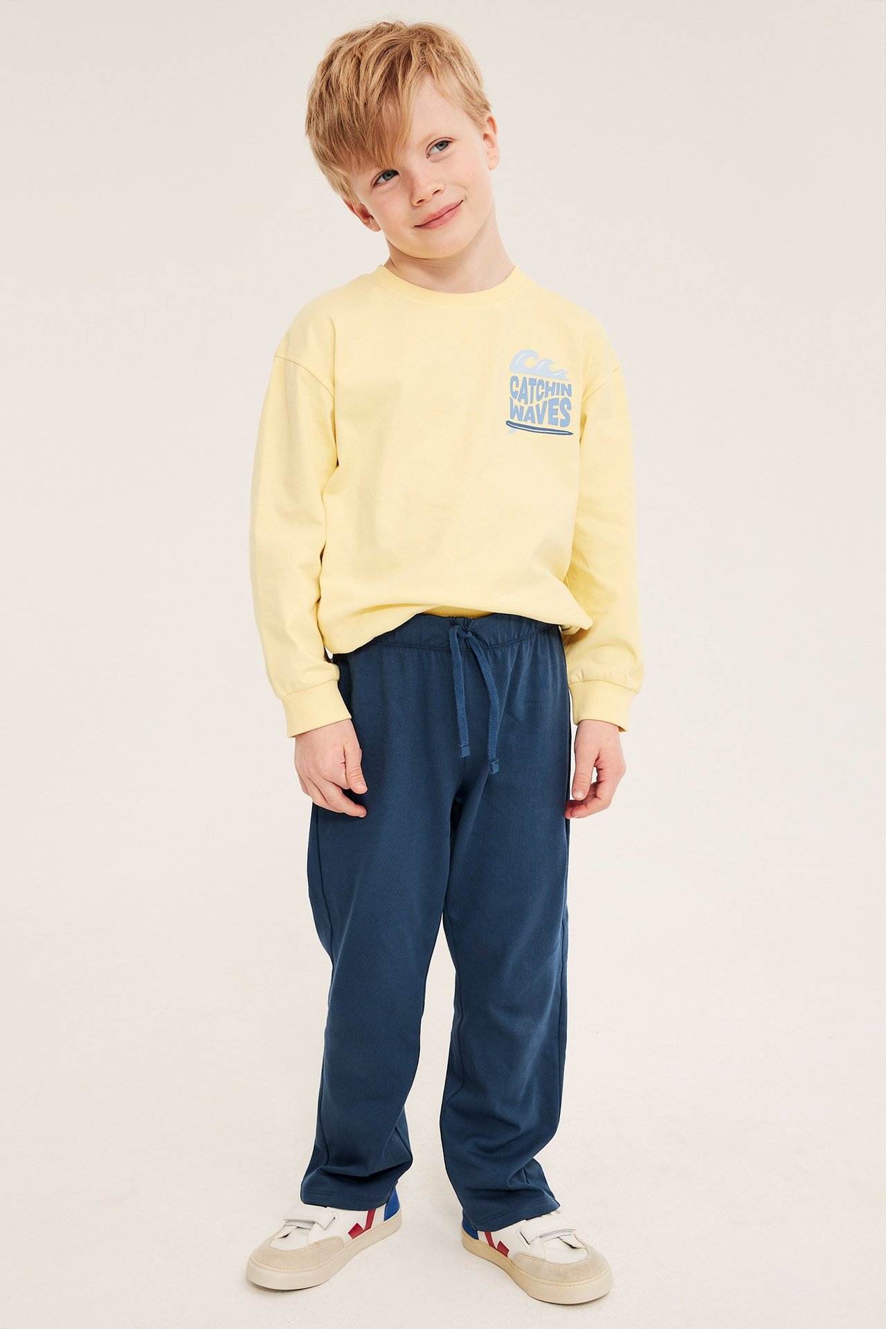 Etunäkymä, lapsi sinisissä collegehousuissa kiristysnyörillä, keltaisessa pitkähihaisessa paidassa ja valkoisissa tennareissa.