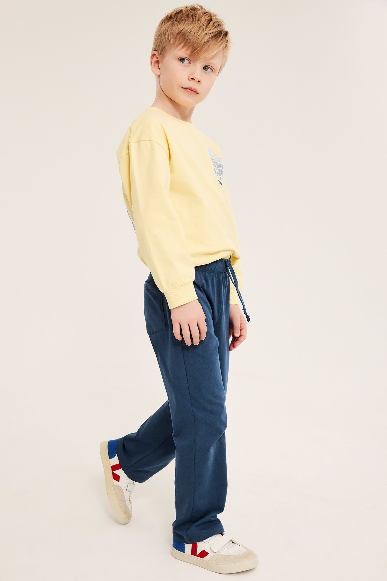 Sivukuva lapsesta, jolla on siniset suoralahkeiset puuvillajoggerit, keltainen pitkähihainen paita ja valkoiset tennarit.
