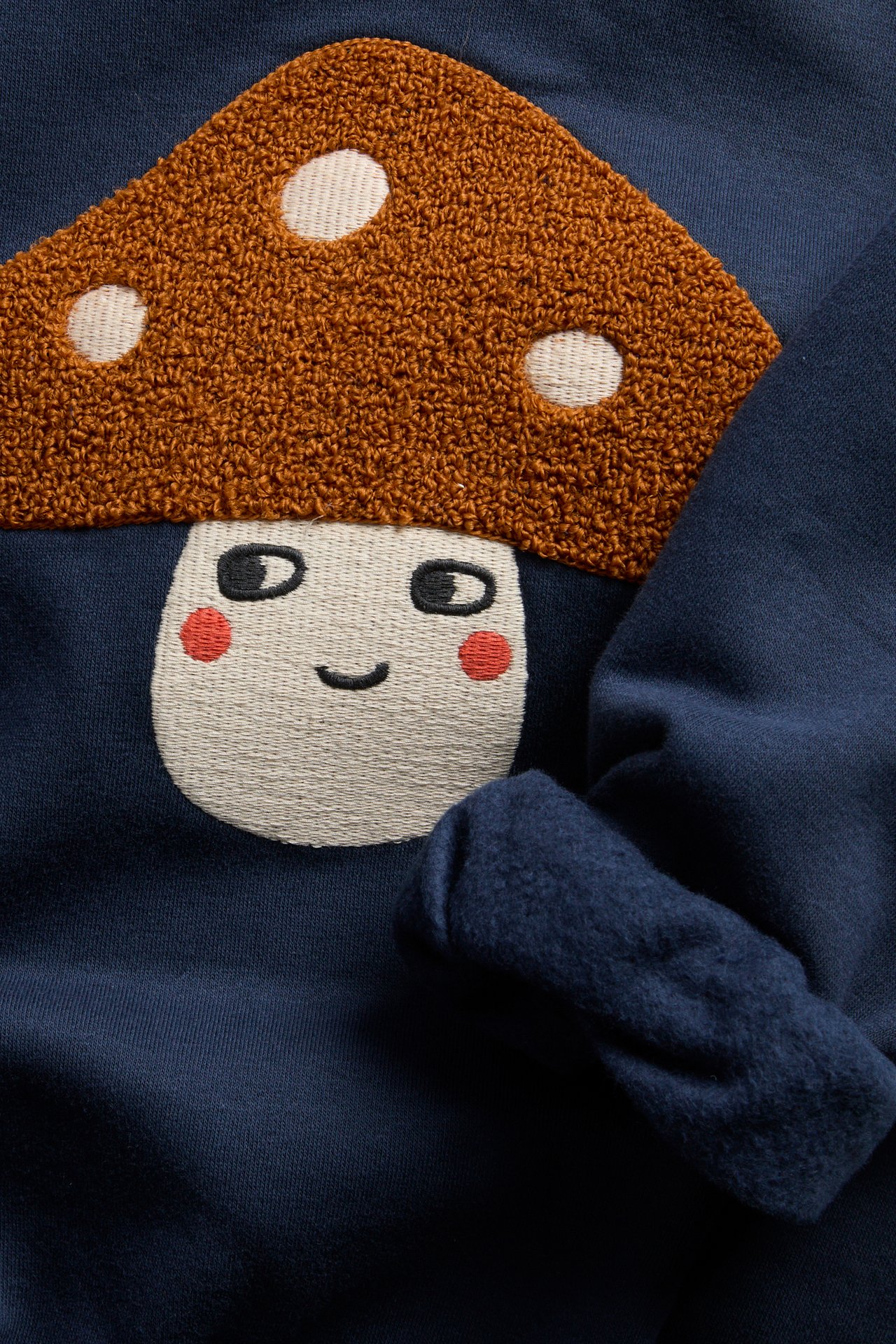 Nærbilde av mørkeblå sweatshirt med teksturert brodert brun sopp og ansikt.
