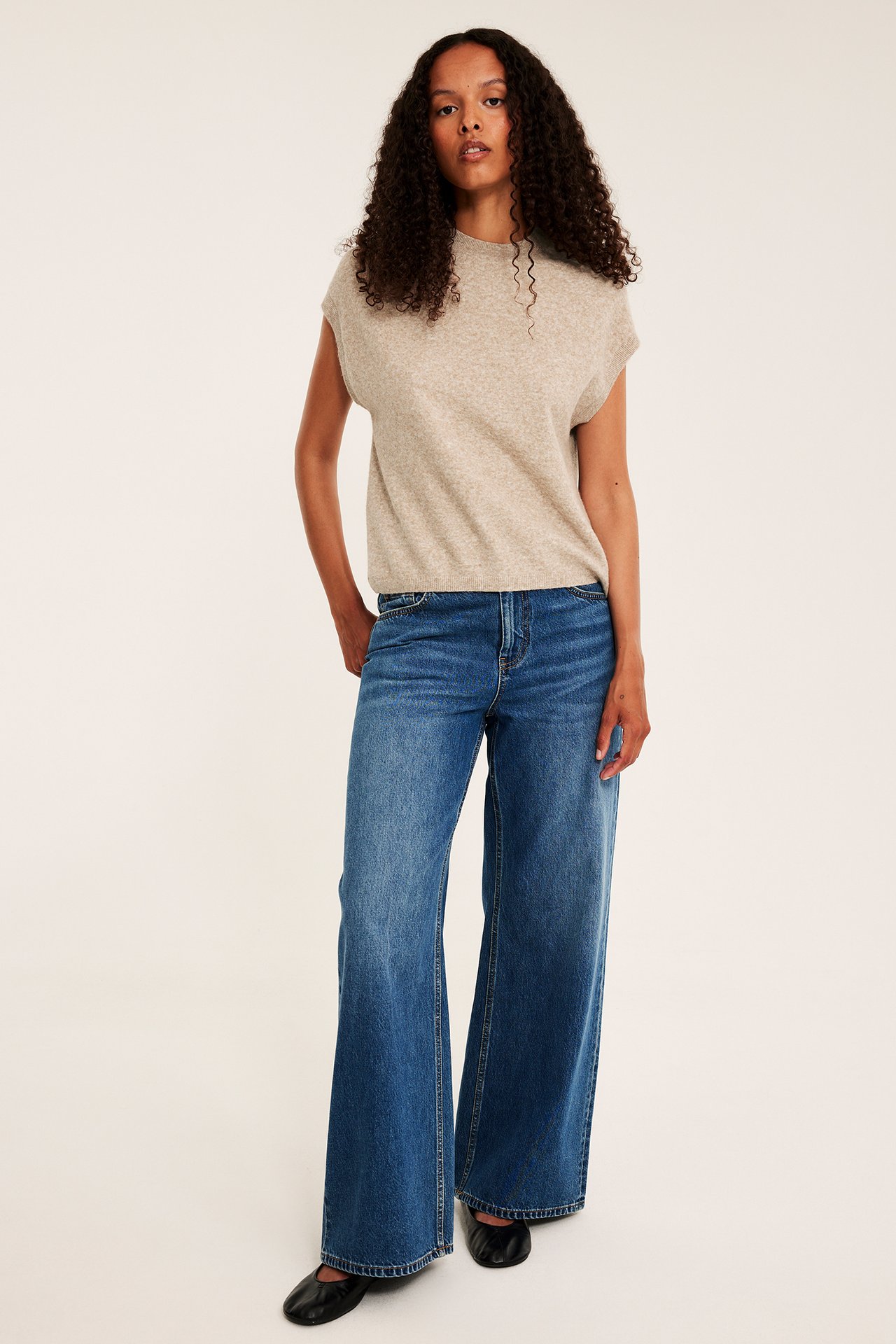 Frontbild på kvinna i beige stickad ärmlös topp, blåa jeans med vida ben och svarta platta skor.