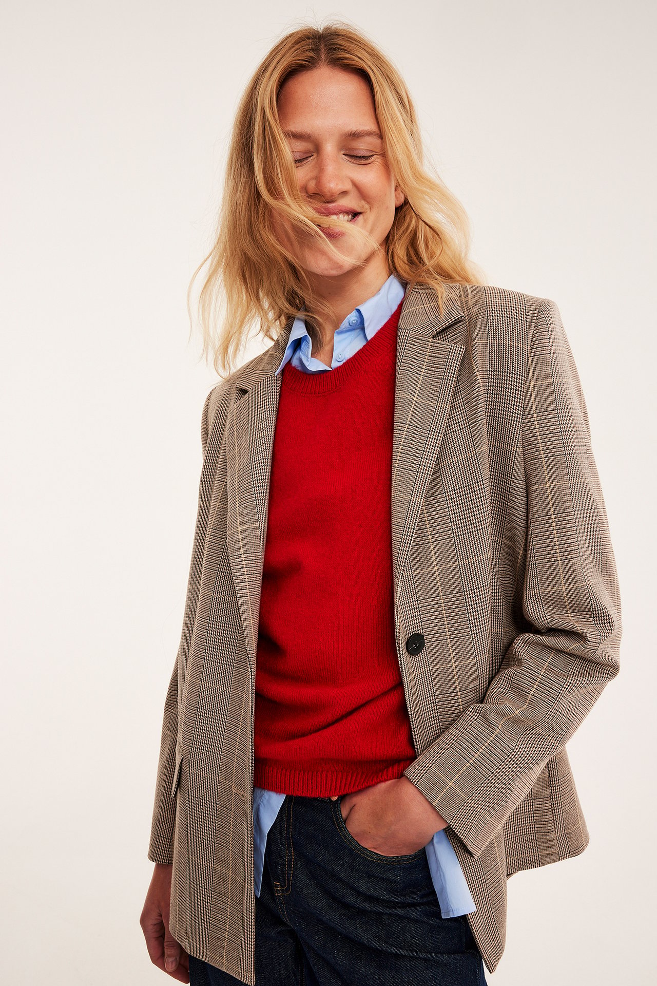 Frontvy av en kvinna i en brunrutig kavaj, röd tröja, blå skjorta och mörkblå jeans.