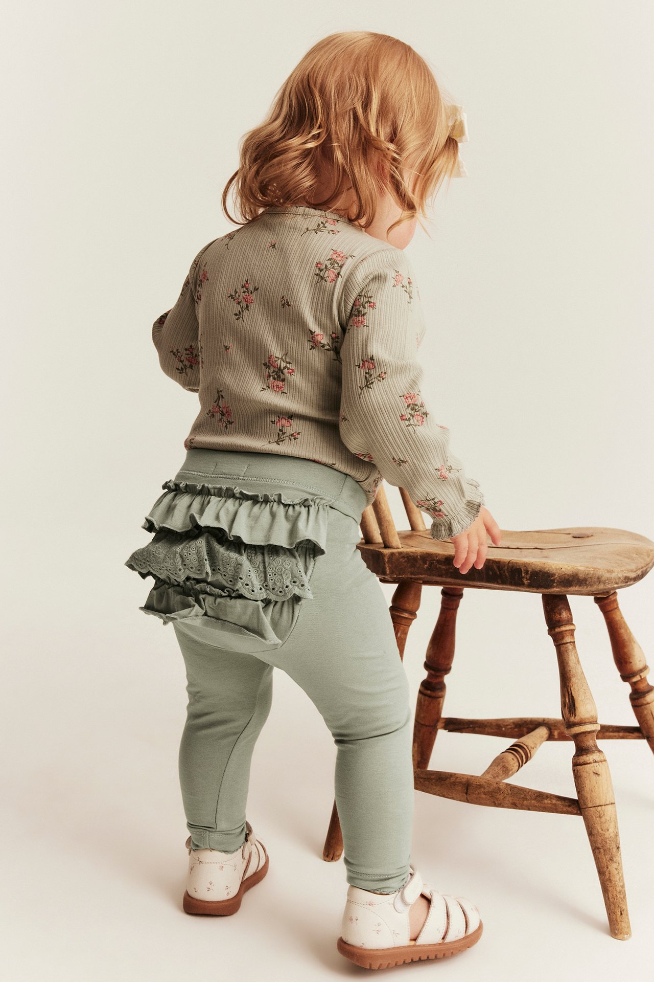 Bakifrån, baby i gröna volang- och spetsleggings, blommig ribbad topp och vita sandaler.