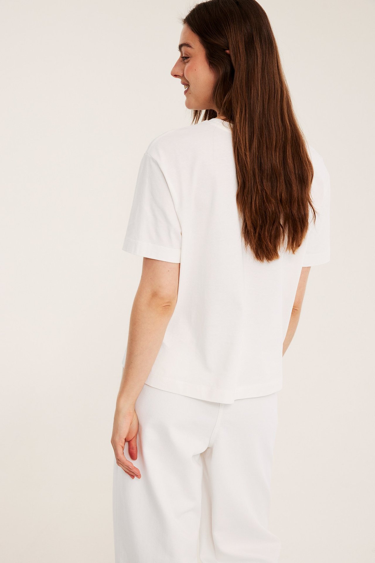 Widok z boku/tyłu kobiety w białym, krótko-rękawowym bawełnianym t-shircie i białych, szerokich spodniach.