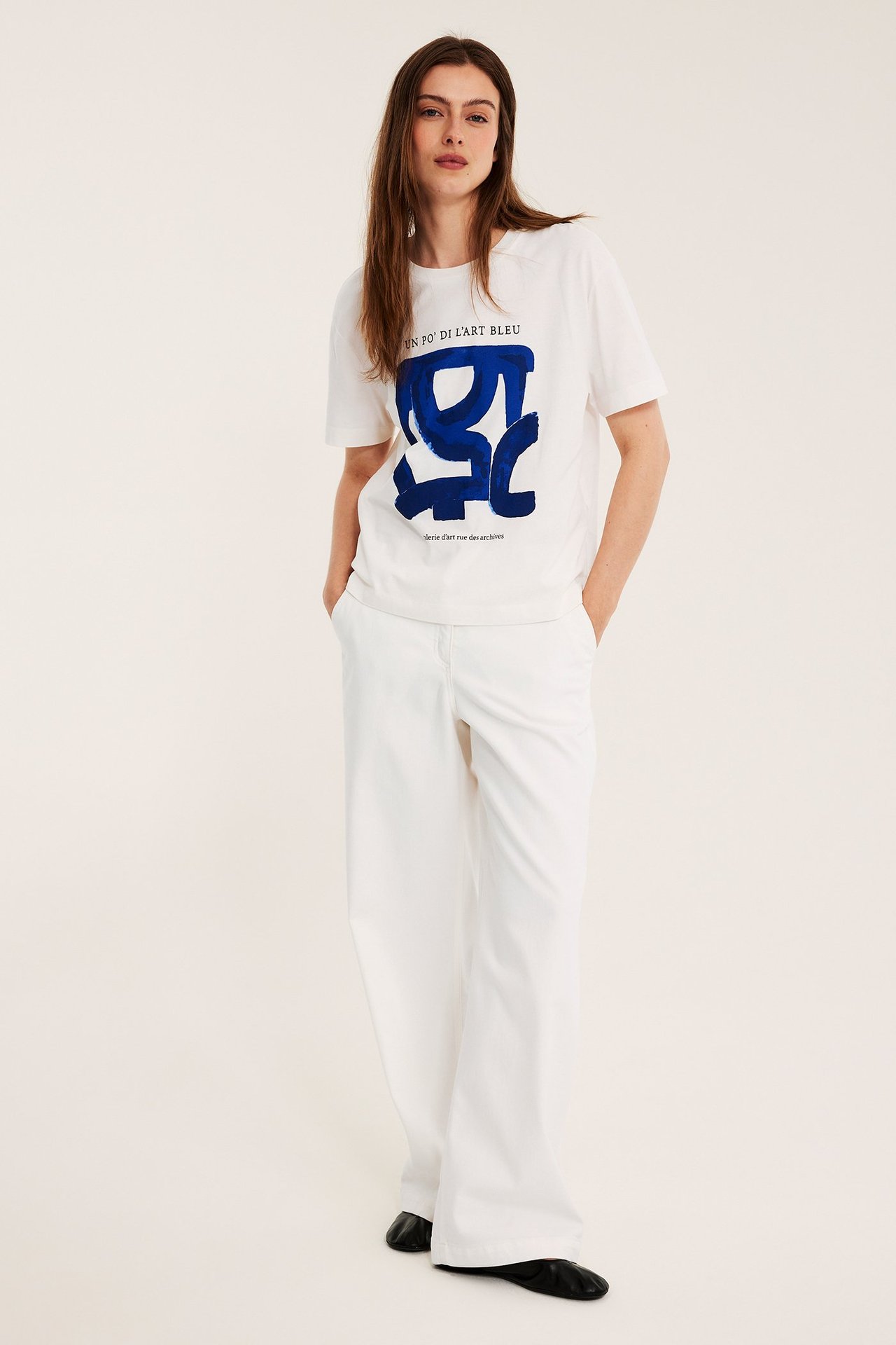 Widok z przodu: kobieta w białym T-shircie z niebieskim nadrukiem, białych szerokich spodniach i czarnych butach.