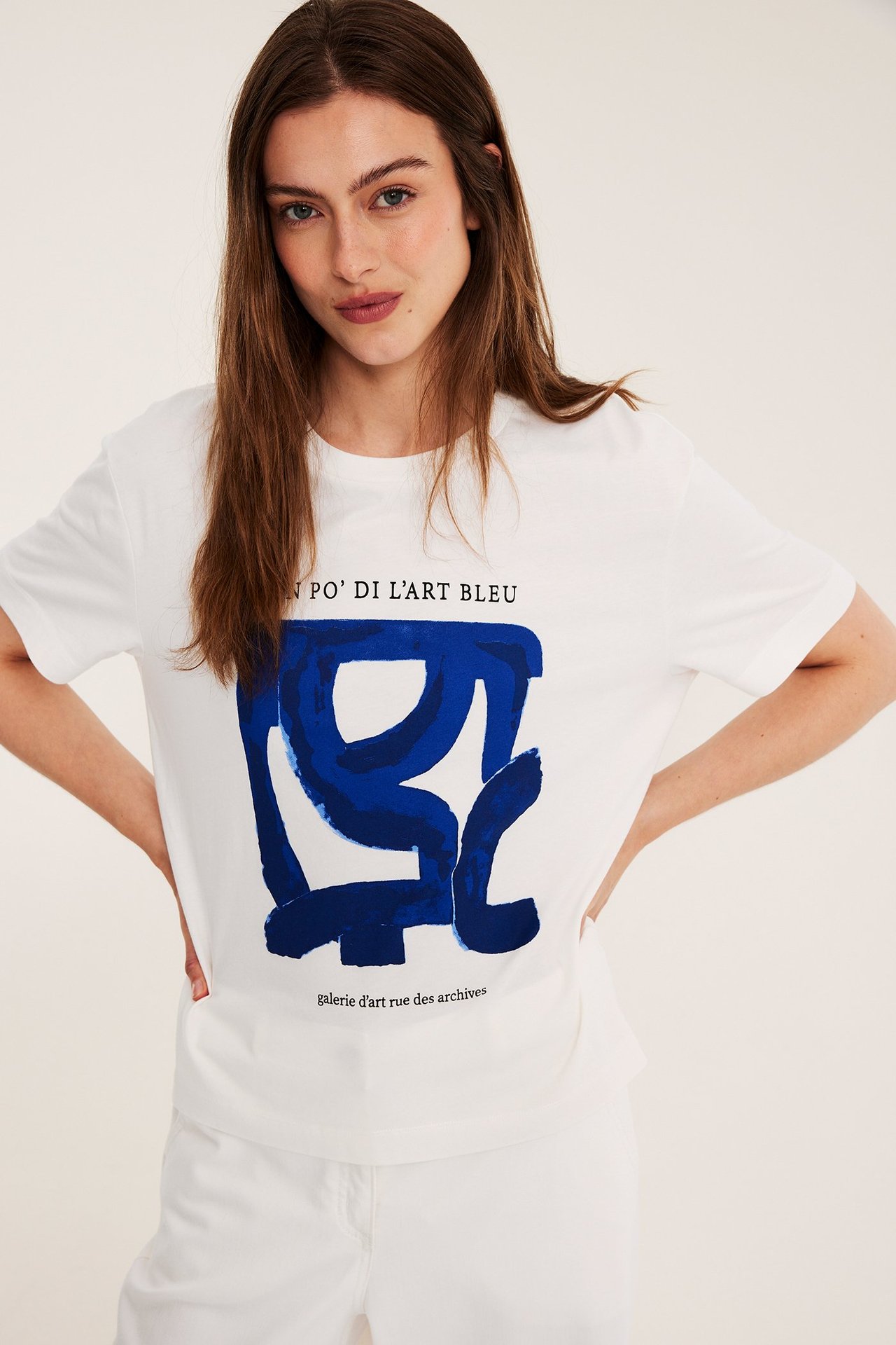 Framifrån: Kvinna i vit bomulls-t-shirt med blått abstrakt tryck, stylad med vita byxor.