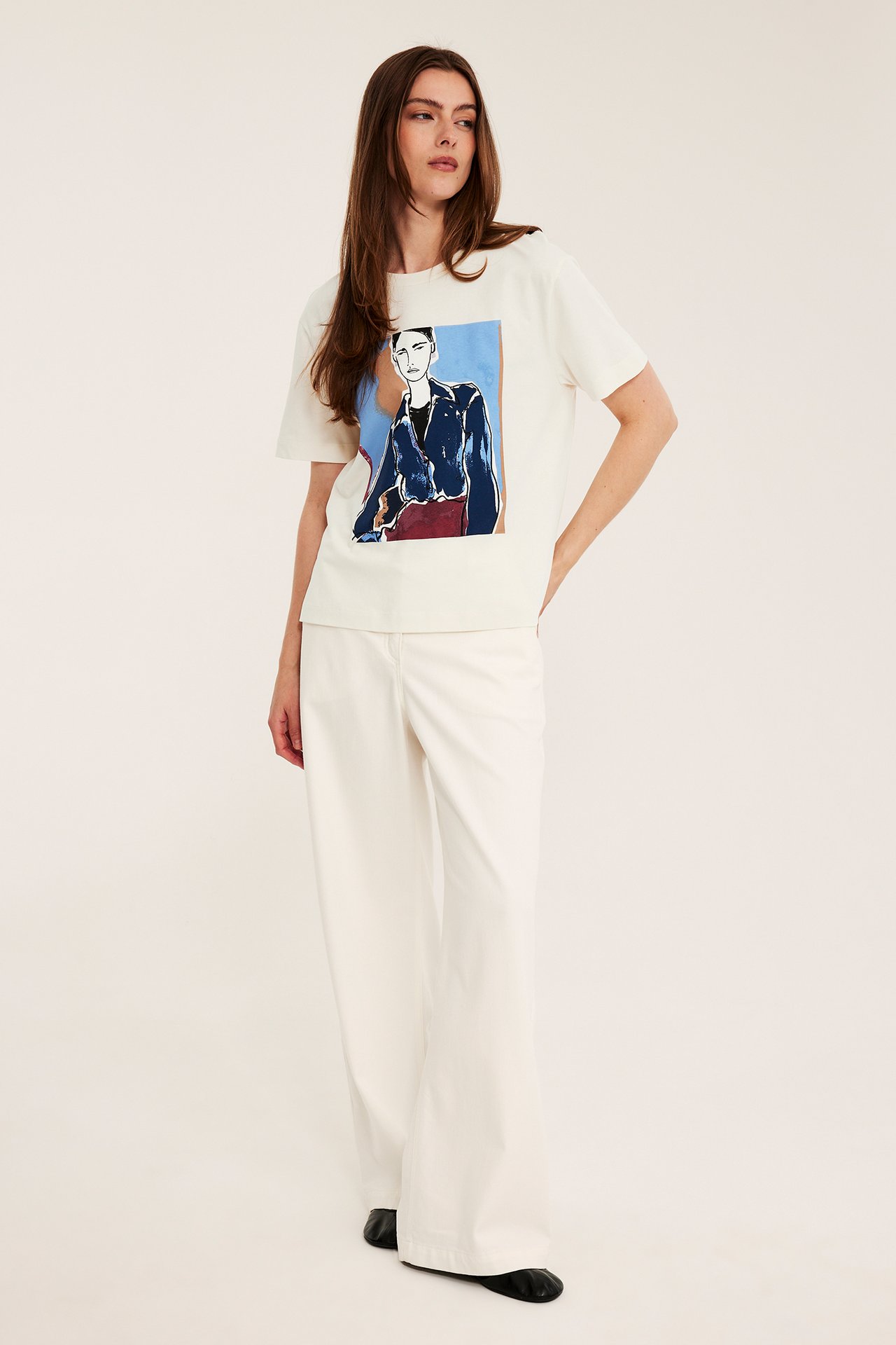 Forfra: kvinne har på seg offwhite bomulls-T-skjorte med fargetrykk av kvinne og offwhite vide bukser.