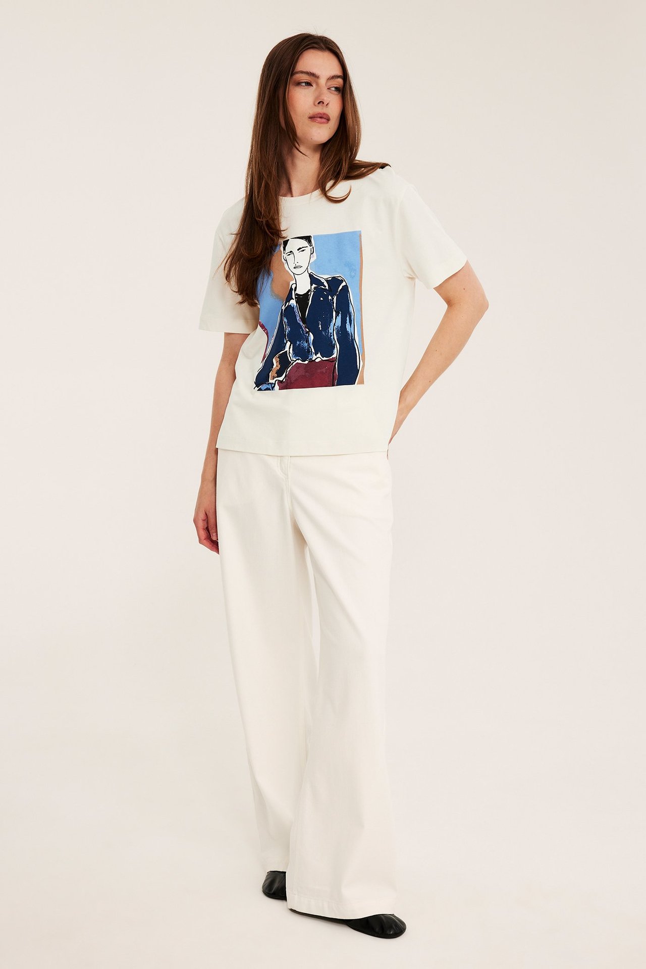 Forfra: kvinne har på seg offwhite bomulls-T-skjorte med fargetrykk av kvinne og offwhite vide bukser.