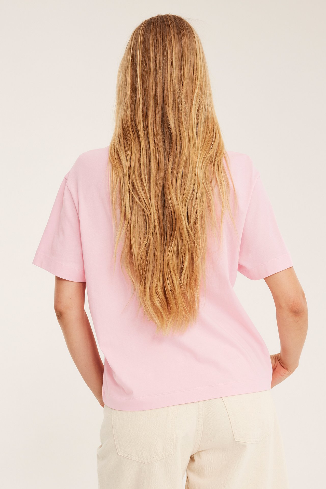 Bakifrån: kvinna i lös rosa kortärmad bomulls-t-shirt och ljusa beigefärgade vida byxor.