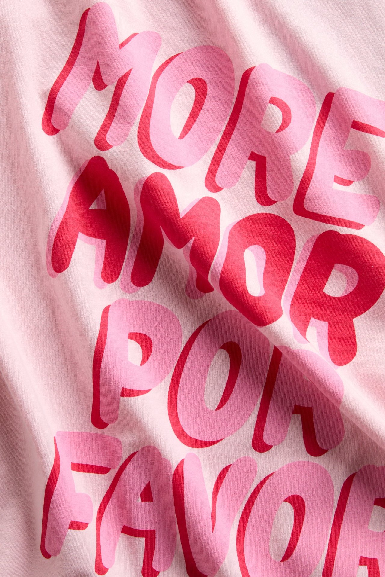 Närbild av en rosa bomulls-t-shirt med rosa och rött tryck "More amor por favor" framtill.