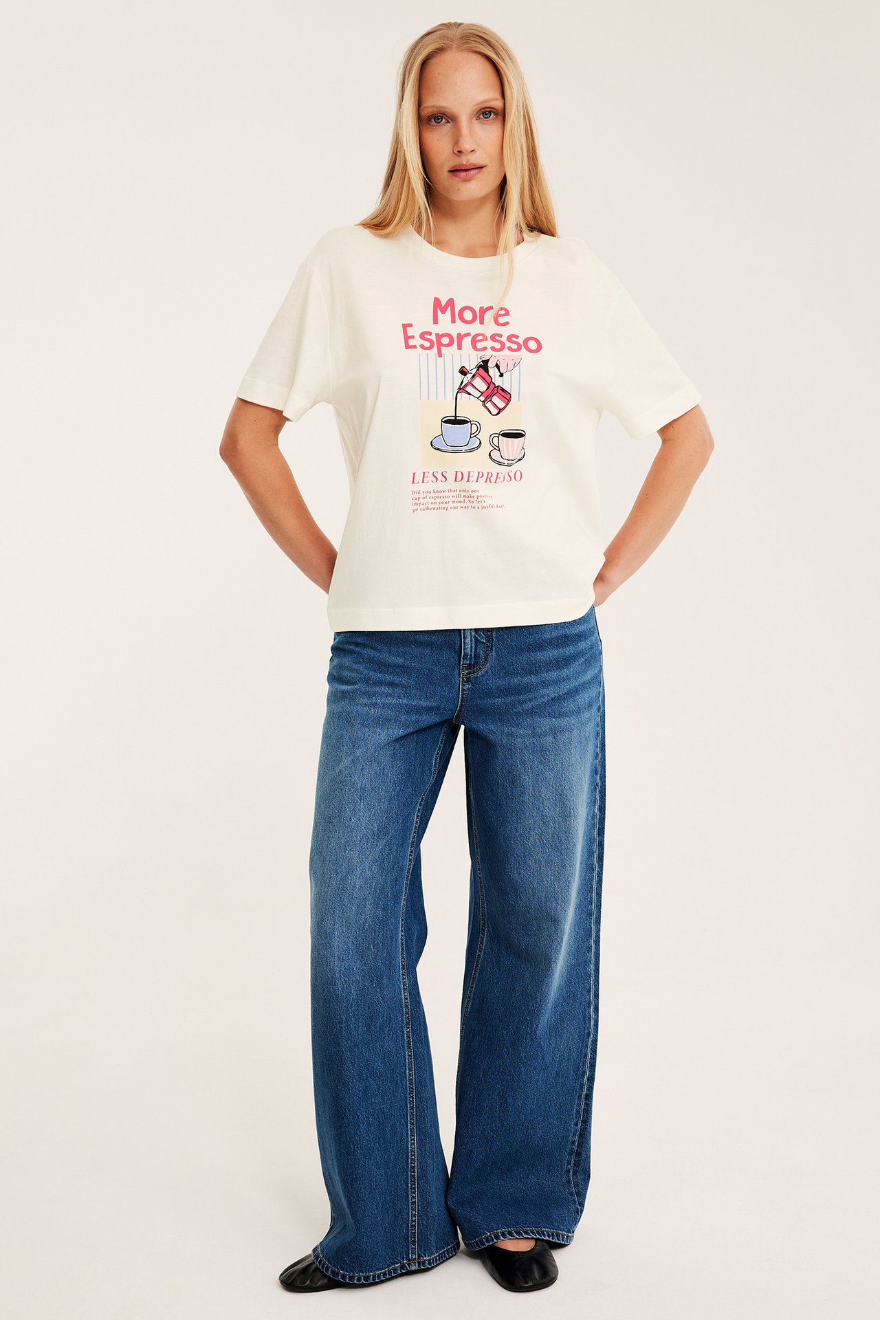 Nainen edestä valkoisessa t-paidassa "More Espresso"-printillä, sinisissä leveälahkeisissa farkuissa ja mustissa kengissä.