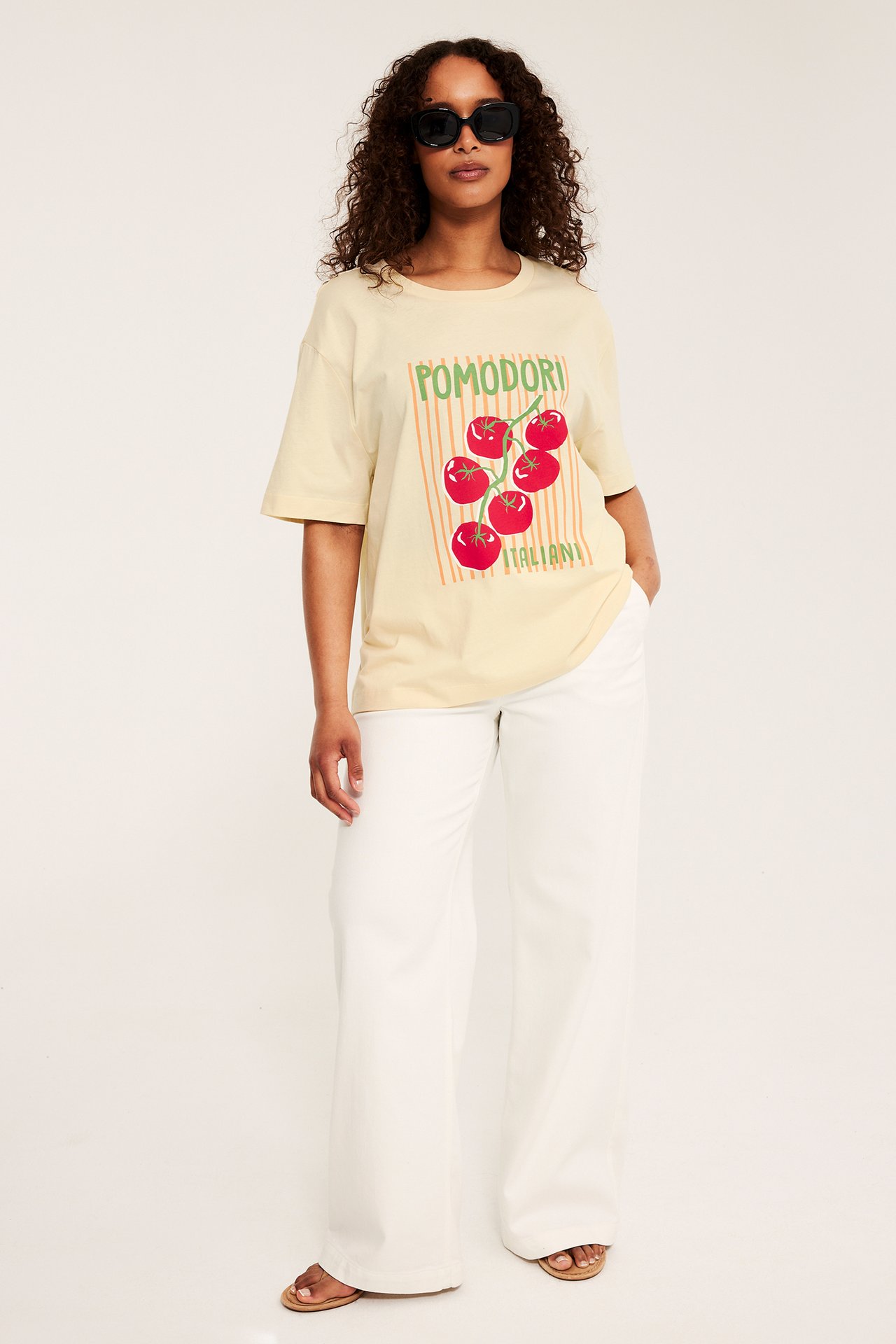 Kvinna bär ljusgul bomulls-t-shirt med tomatmotiv, vita vida byxor, sandaler och svarta solglasögon, framifrån.