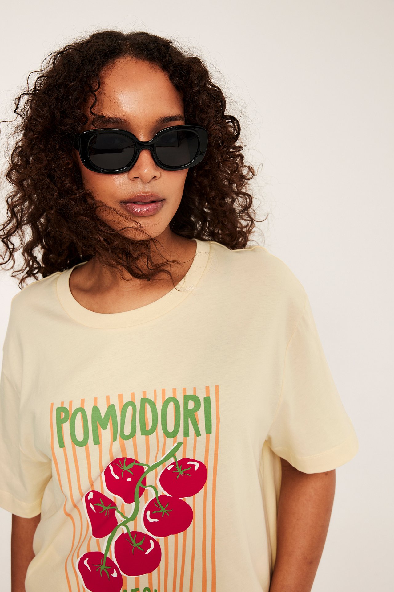 Närbild: Ljusgul bomulls-t-shirt med 'Pomodori'-tryck på kvinna i svarta solglasögon.