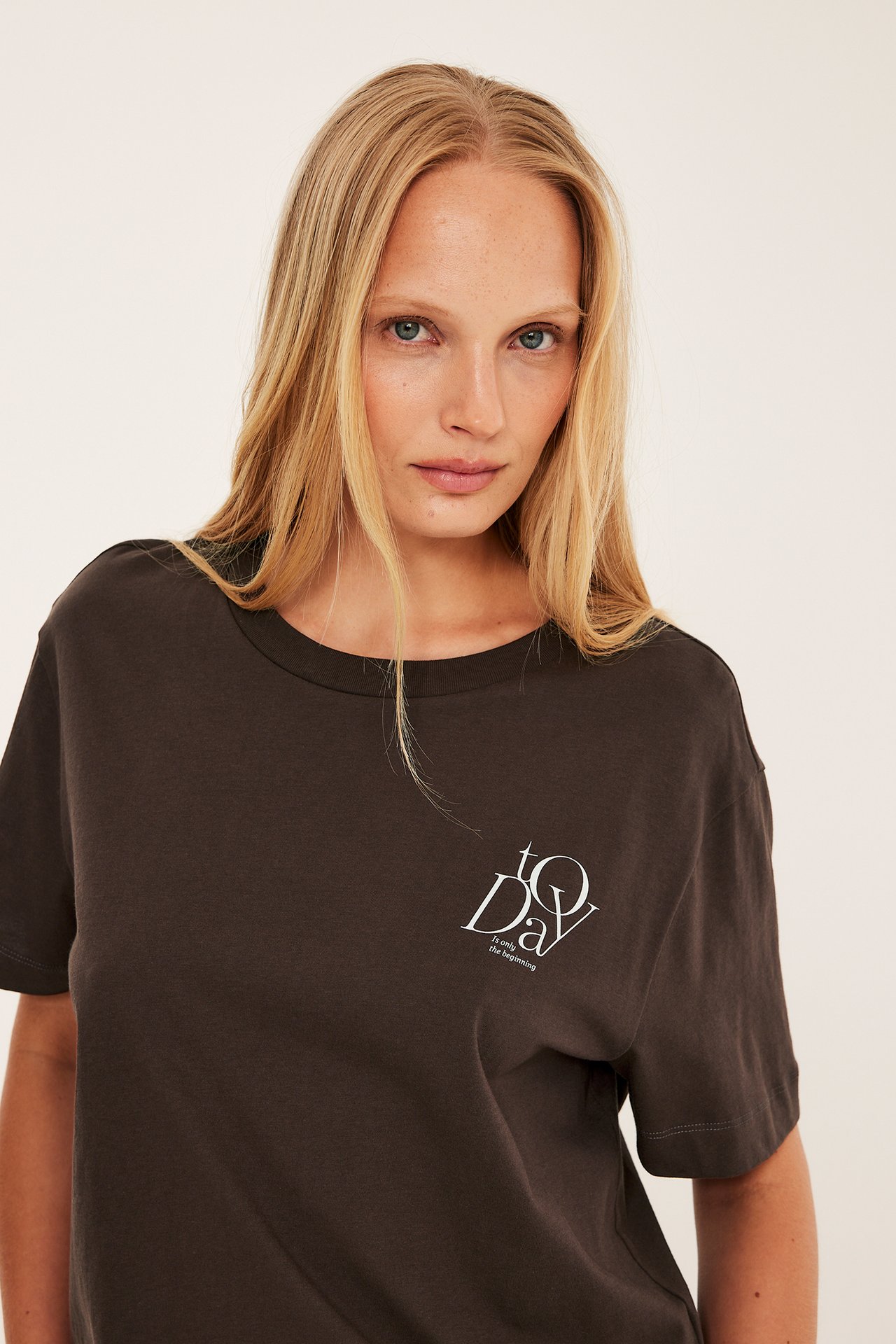 Närbild framifrån på en kvinna i en mörkbrun bomulls-t-shirt med vitt "TODAY"-brösttryck.